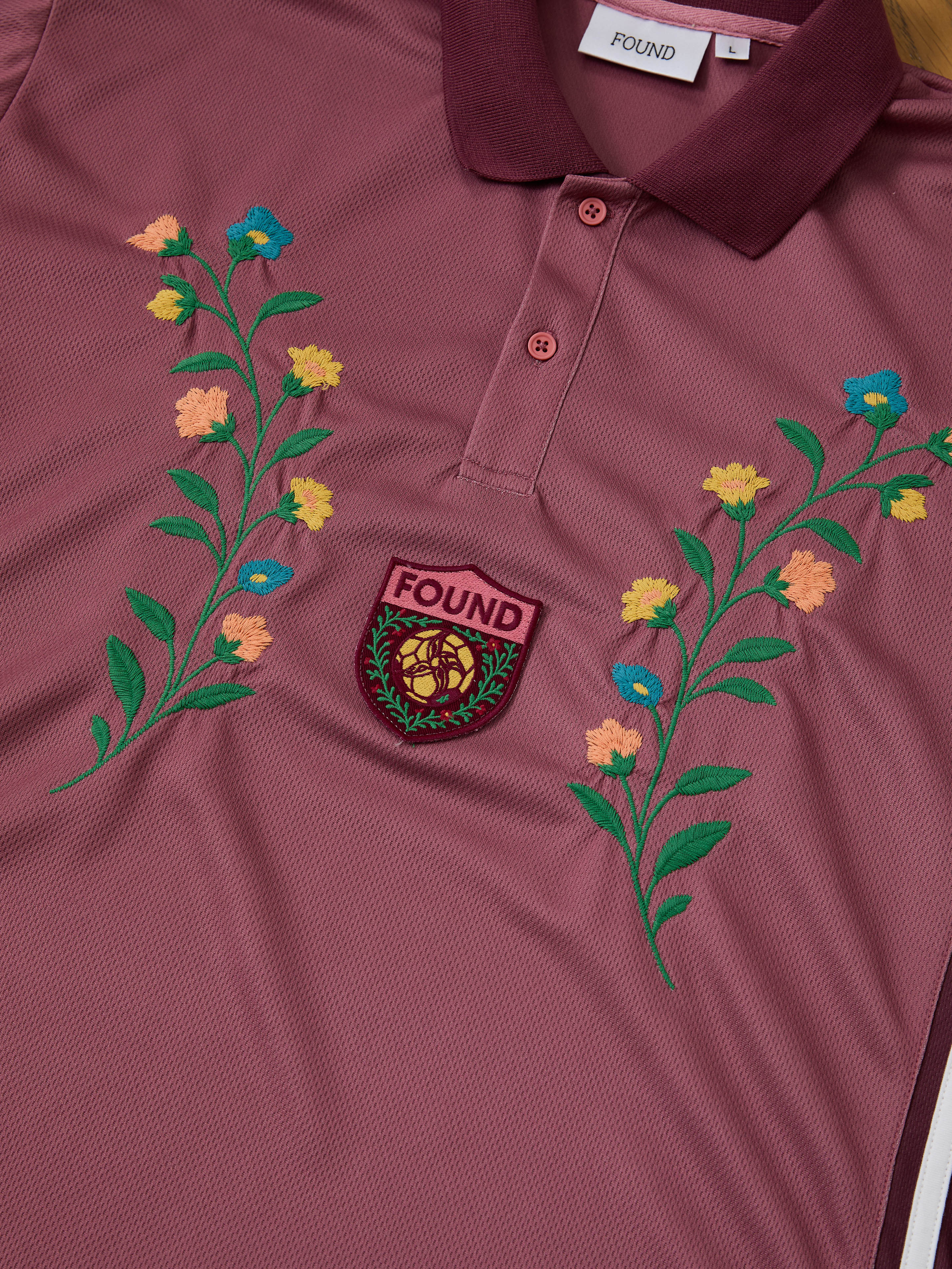 The FOUND Floral Polo Jersey features a maroon cotton build, floral embroidery on the chest, and a FOUND crest patch—an elegant take on classic vintage football shirt style.