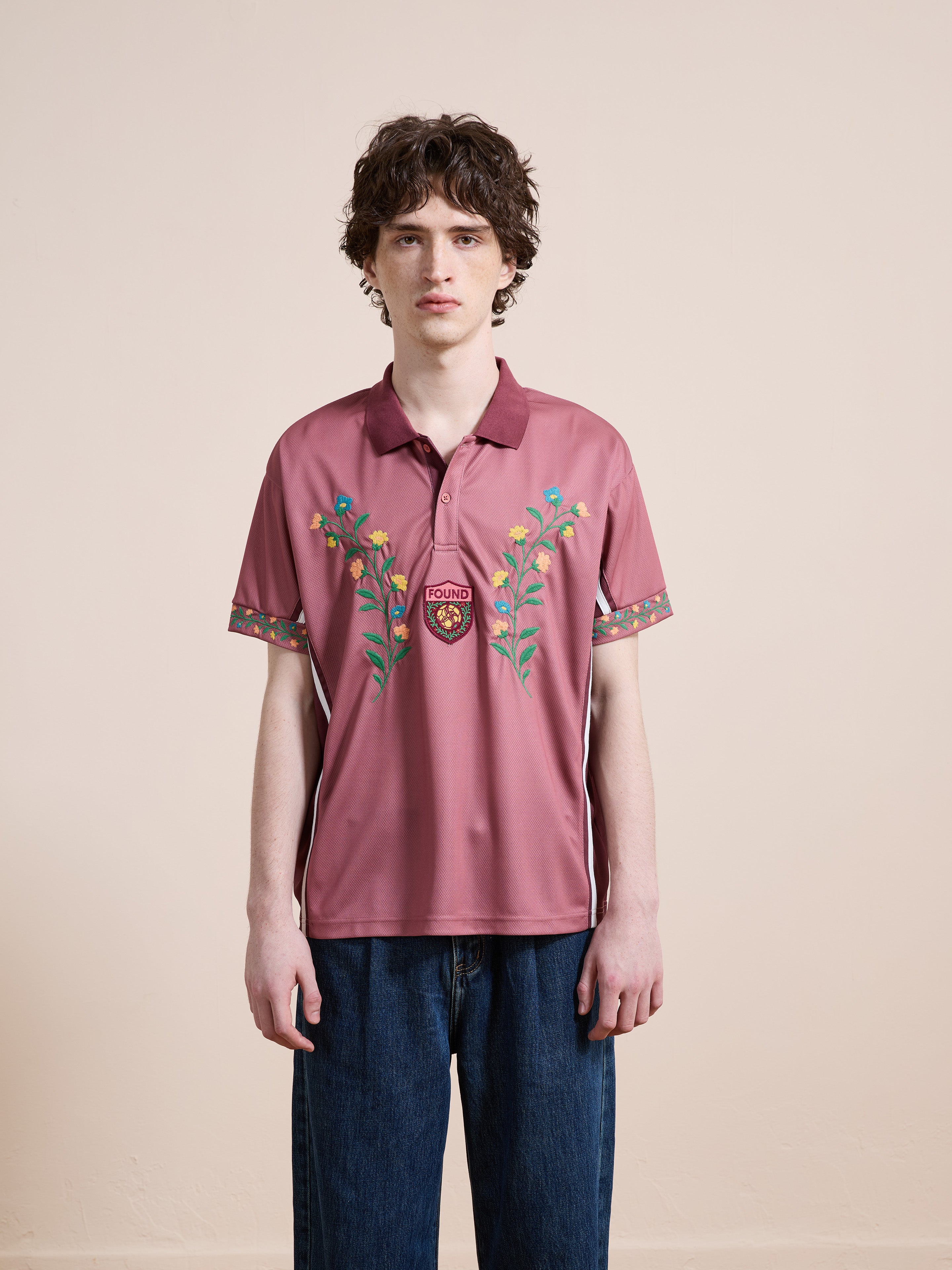 A young man with curly hair wears the FOUND Floral Polo Jersey and blue jeans, standing against a plain beige background.