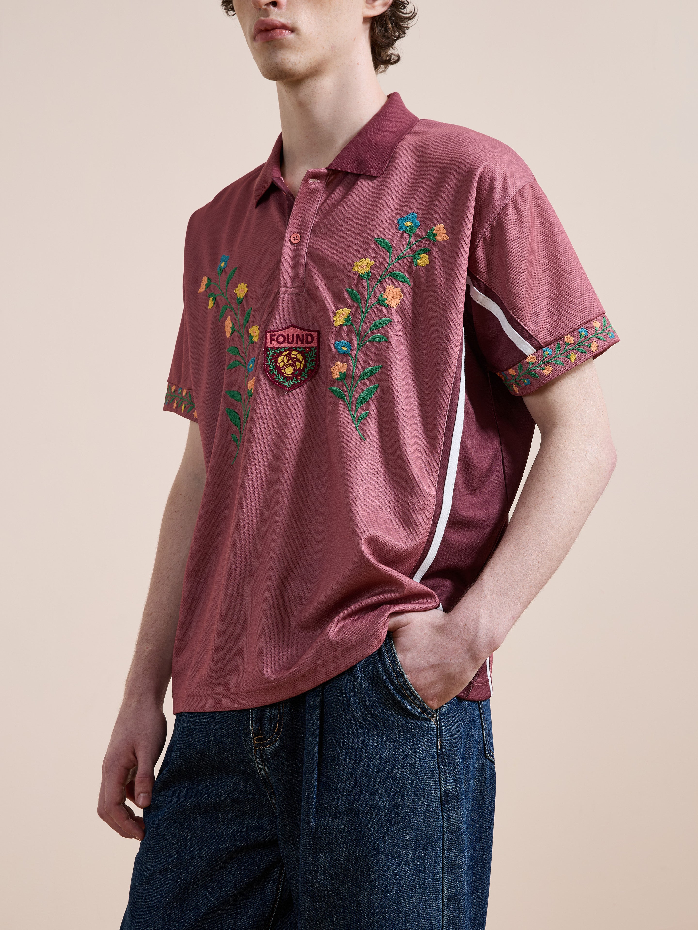 A person wears the FOUND Floral Polo Jersey—a maroon cotton polo with floral embroidery—paired with blue jeans, standing against a neutral background.