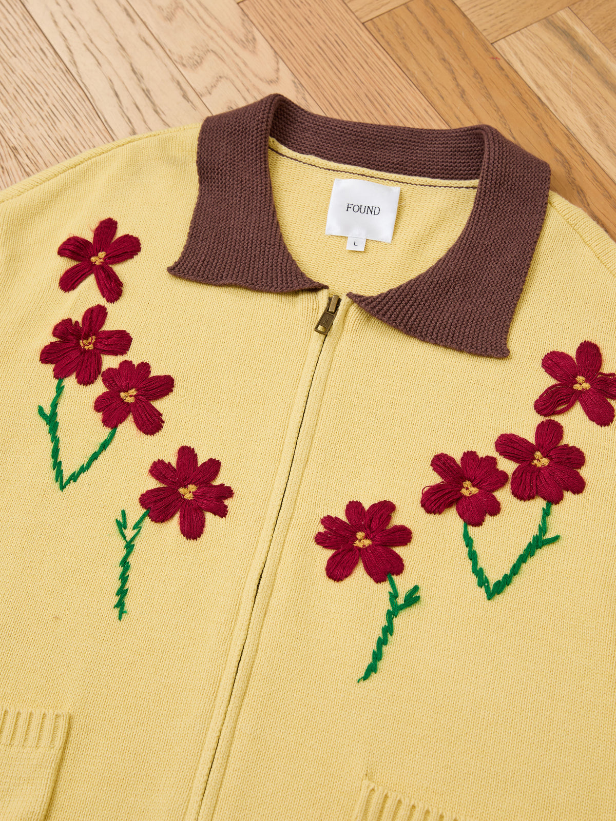 The FOUND Floral Sun Knit Sweater, featuring yellow knit with a brown collar and red floral embroidery, is laid flat on a wooden floor.