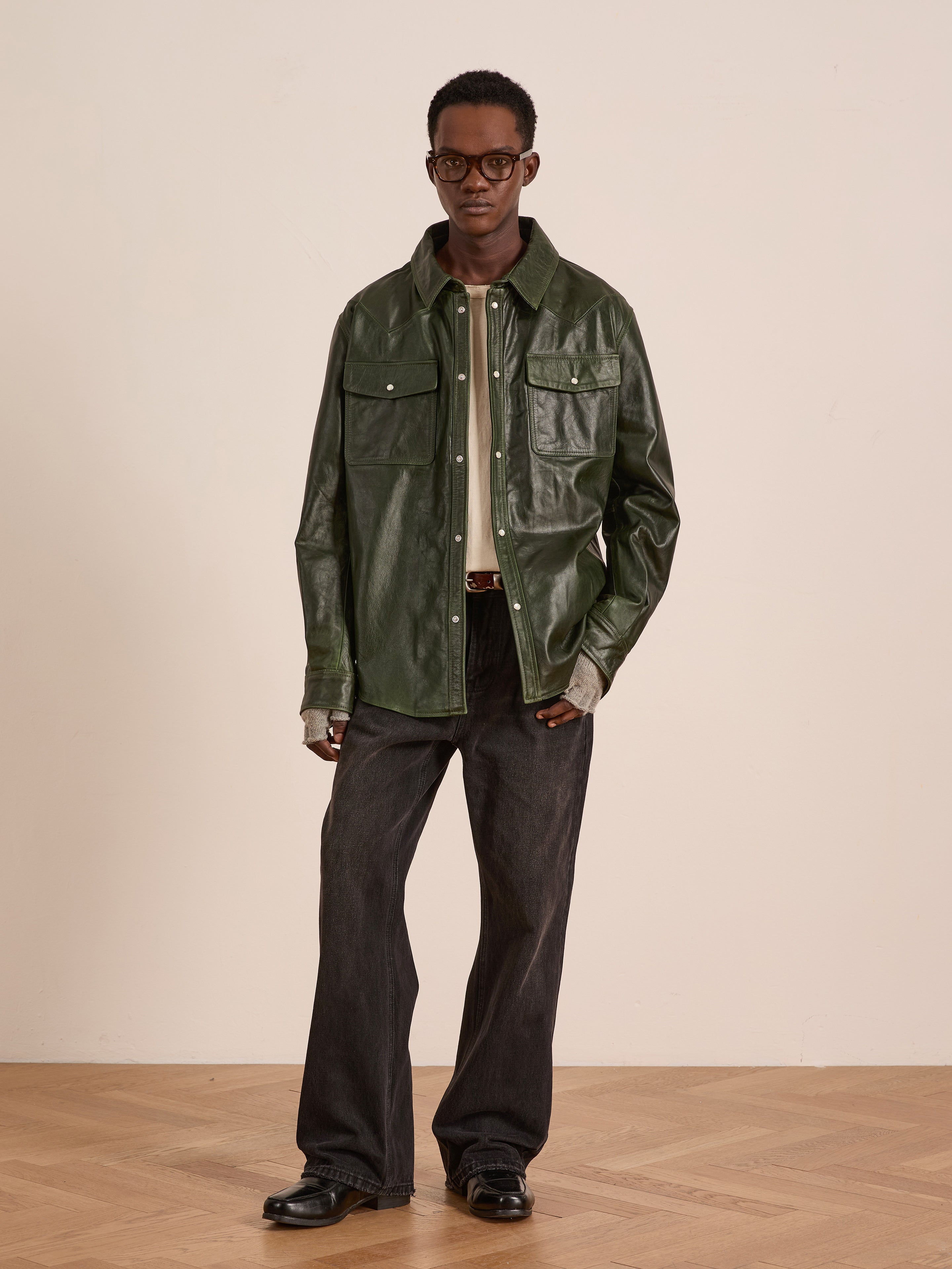 A man stands indoors against a light wall, wearing FOUND’s Forest Leather Western Shirt paired with dark jeans, black shoes, and glasses.