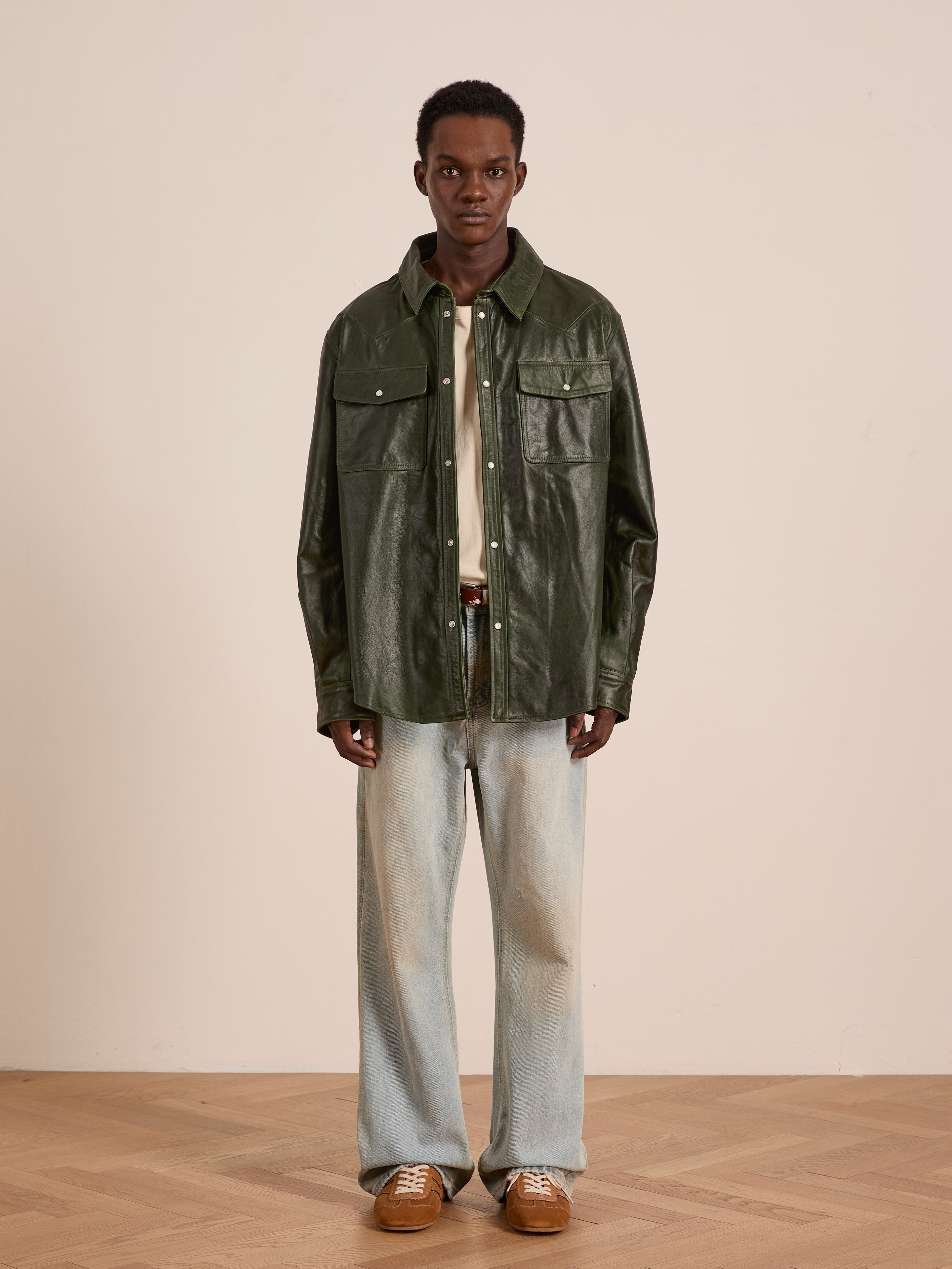 Indoors, a person wears the FOUND Forest Leather Western Shirt layered over a beige shirt, paired with light blue wide-leg jeans and brown sneakers.