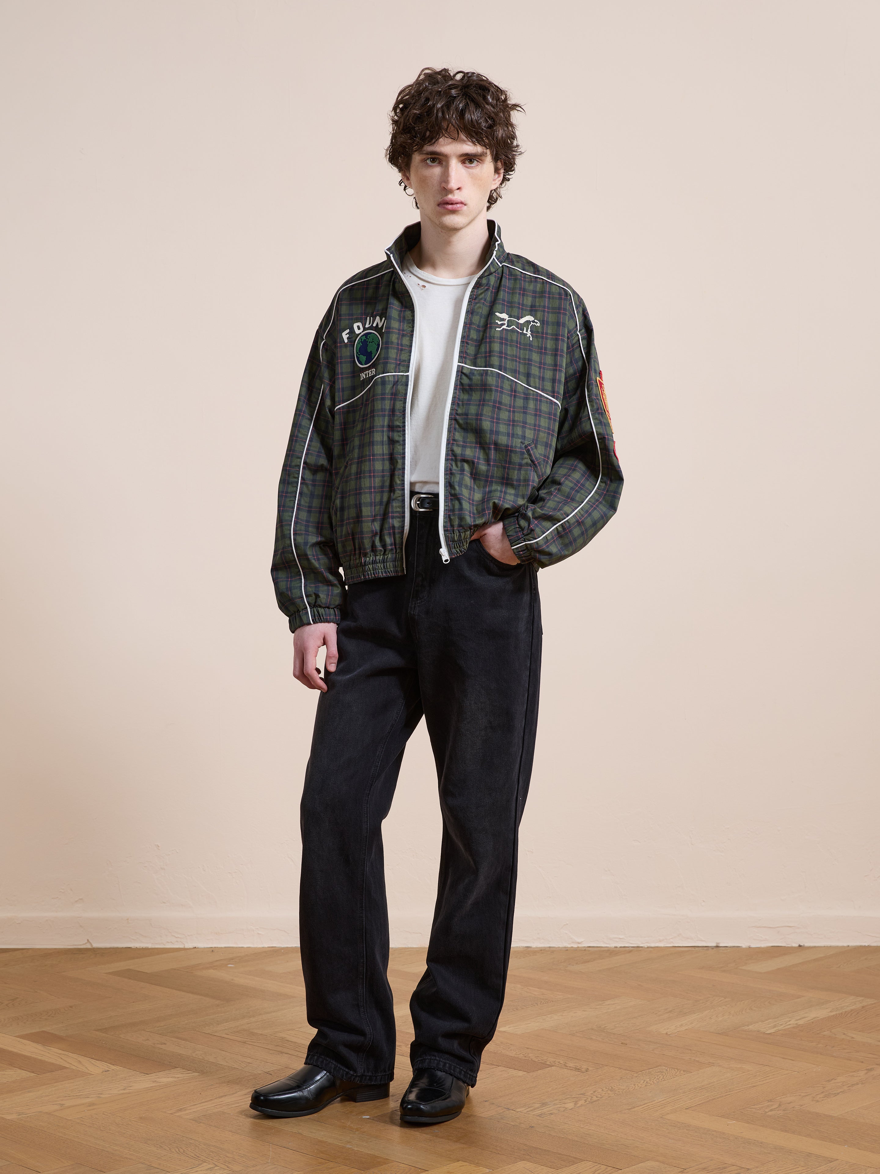 Indoors on a wooden floor, a person wears the FOUND Forest Plaid Track Jacket over a white t-shirt, paired with black pants and black shoes, standing against a plain beige wall.