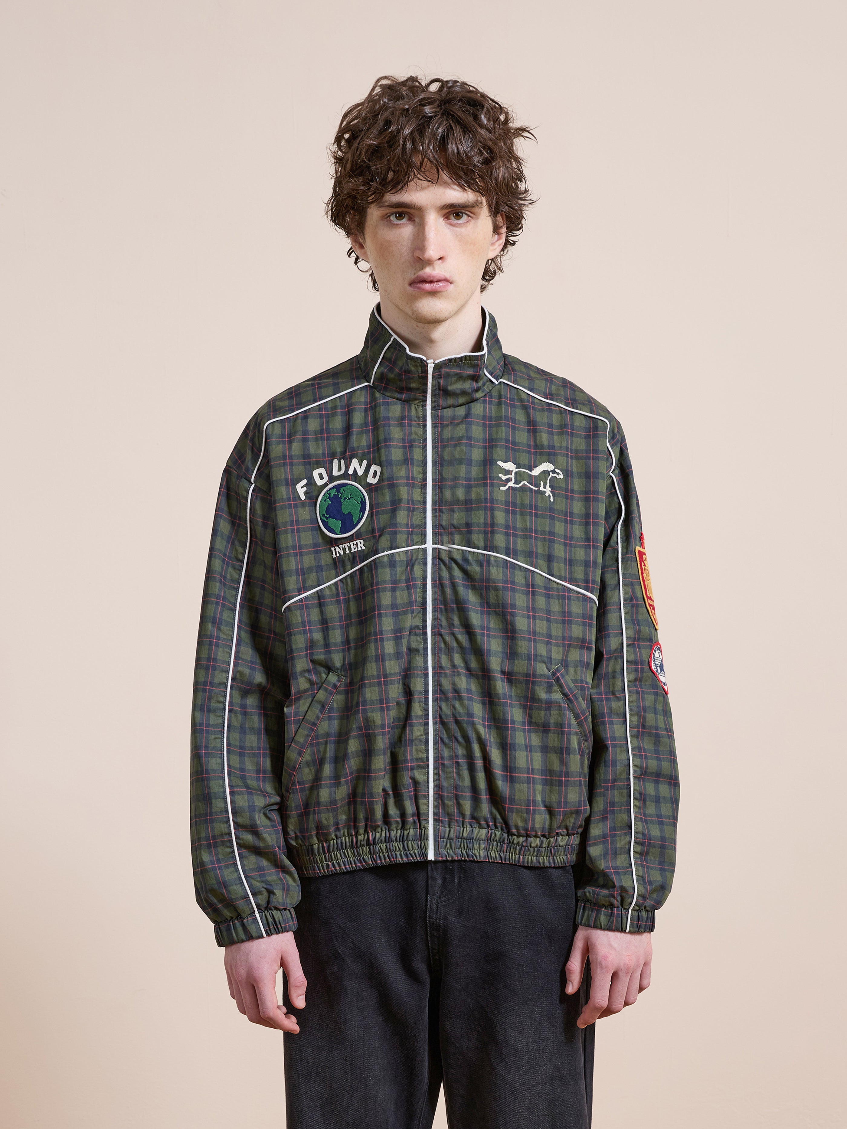 A curly-haired person wears the FOUND Forest Plaid Track Jacket, featuring lightweight cotton and patches, while standing against a plain beige background.
