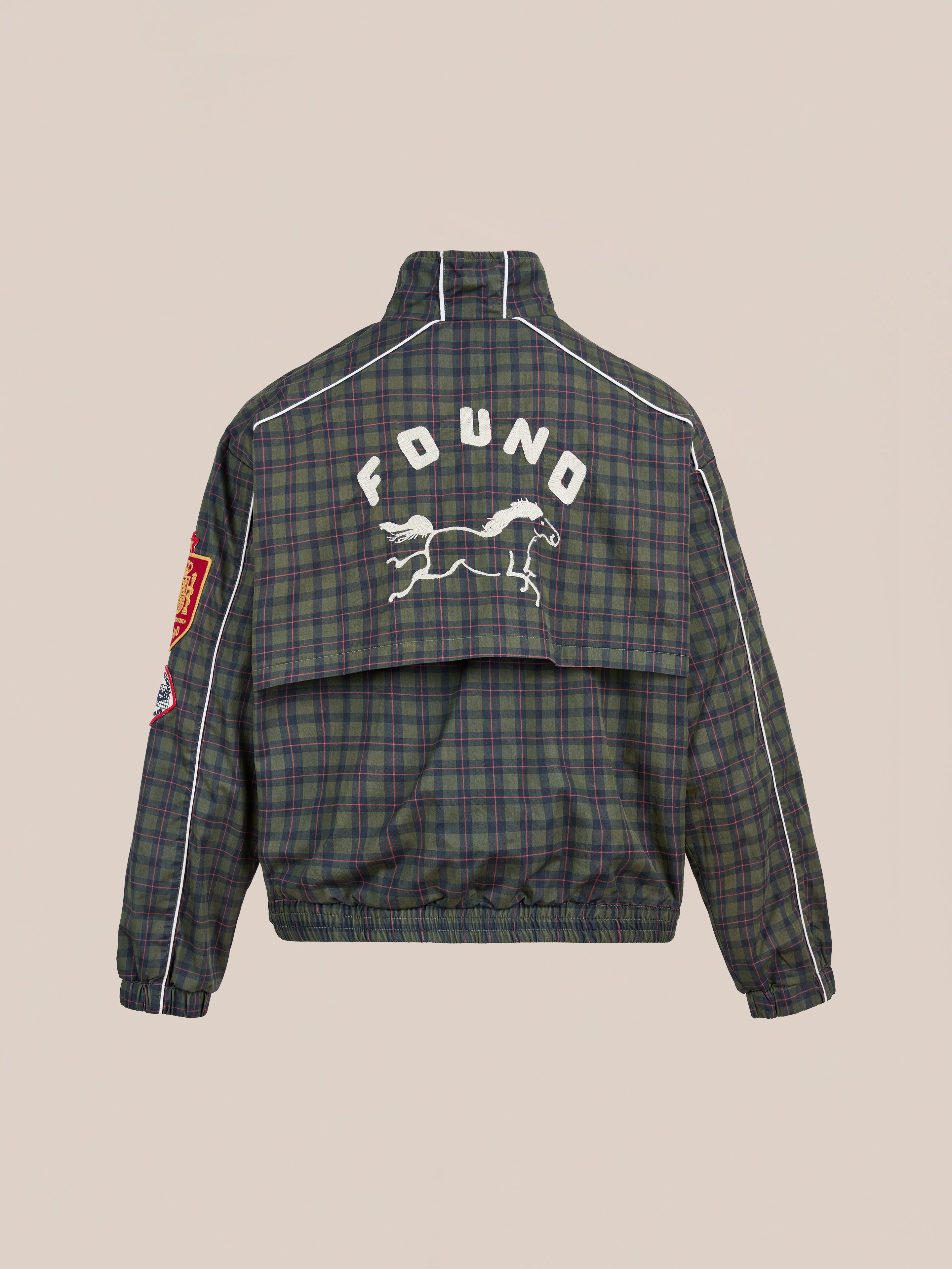 The FOUND Forest Plaid Track Jacket features green and navy plaid with white piping, a red sleeve patch, and a leaping animal graphic plus "FOUND" on the back—a lightweight cotton jacket inspired by vintage sport style.
