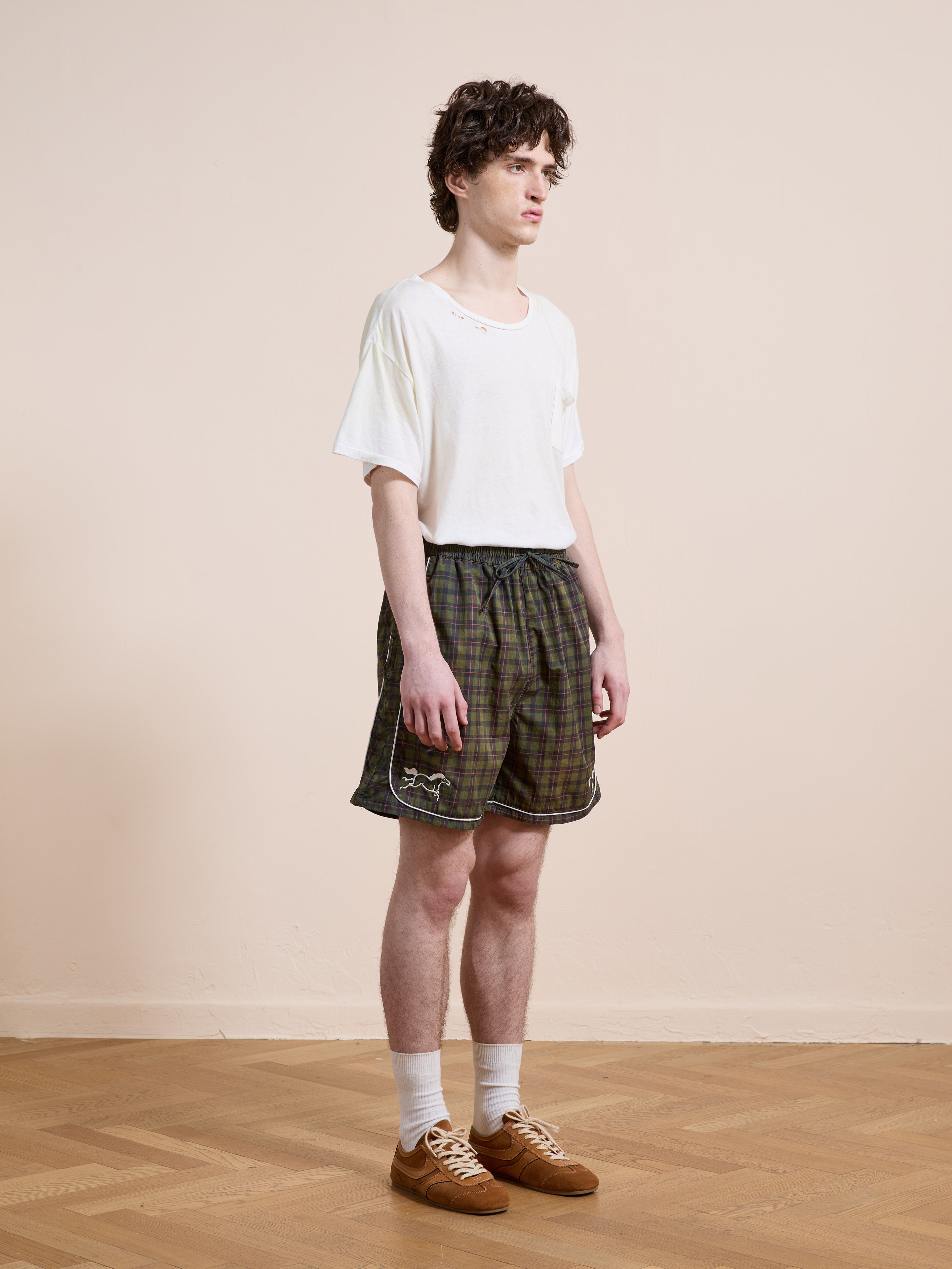 A young man stands indoors on a wooden floor against a beige wall, wearing Found's Forest Plaid Track Shorts with a white t-shirt, white socks, and brown shoes.