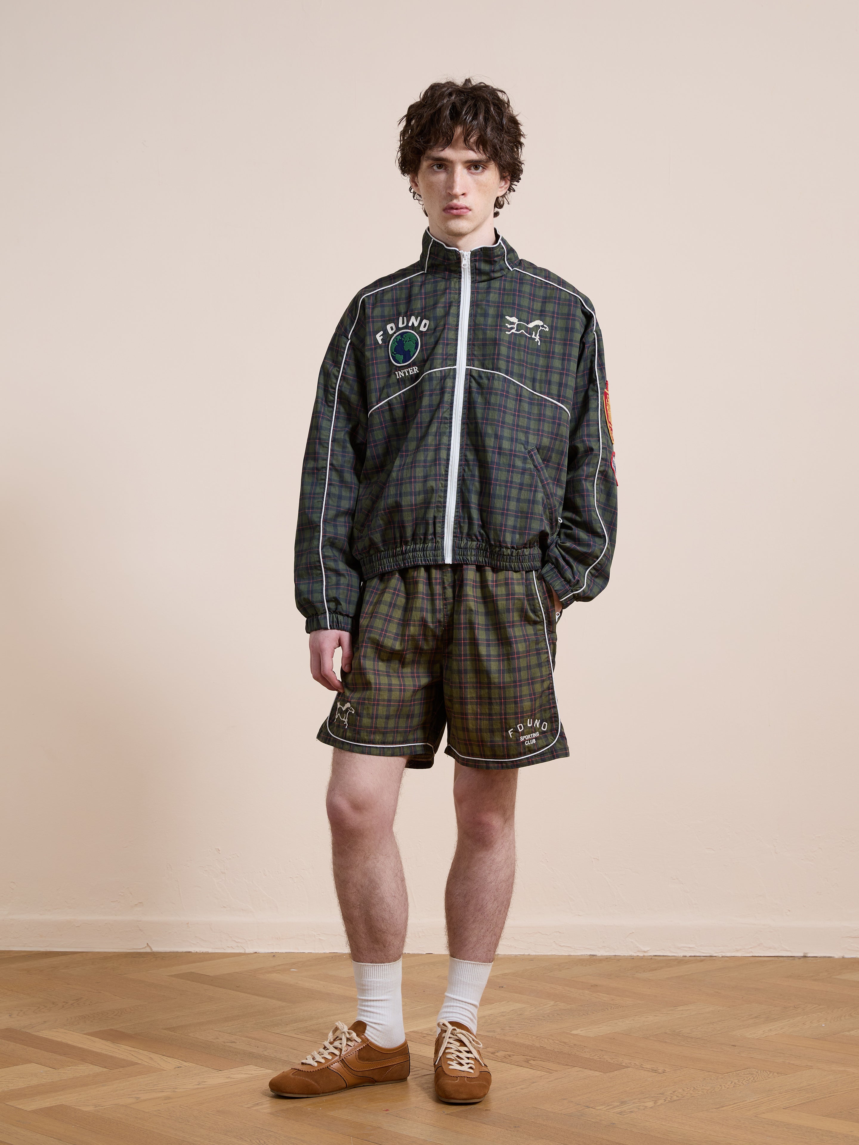 A person stands indoors in a matching plaid track jacket and Found’s Forest Plaid Track Shorts, highlighting the unisex relaxed fit. White socks and brown sneakers complete the vintage athletic look on a wooden floor against a beige wall.