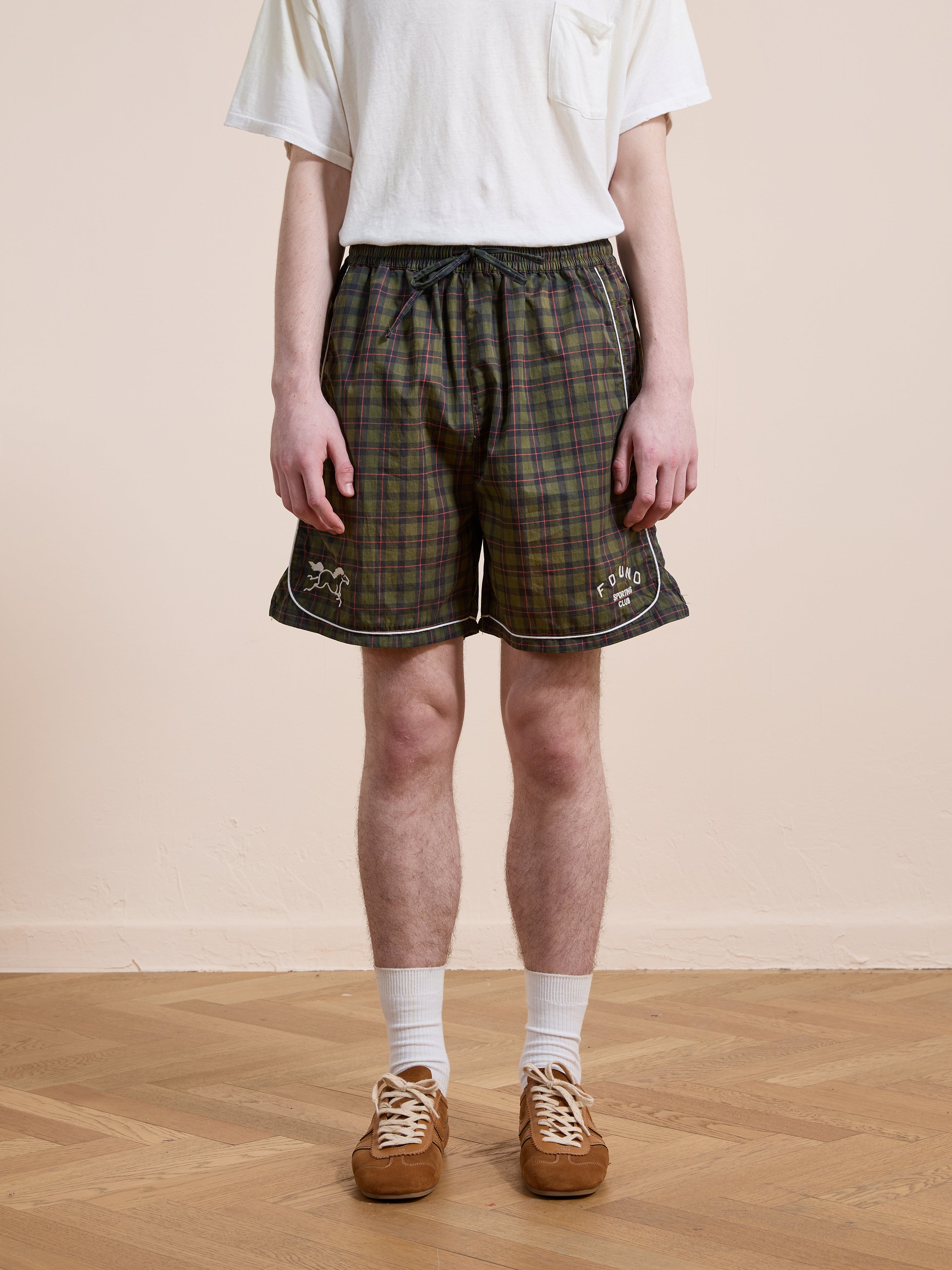 A person stands on a wooden floor against a beige wall, wearing a white T-shirt, Found Forest Plaid Track Shorts (unisex relaxed fit), white socks, and brown sneakers.