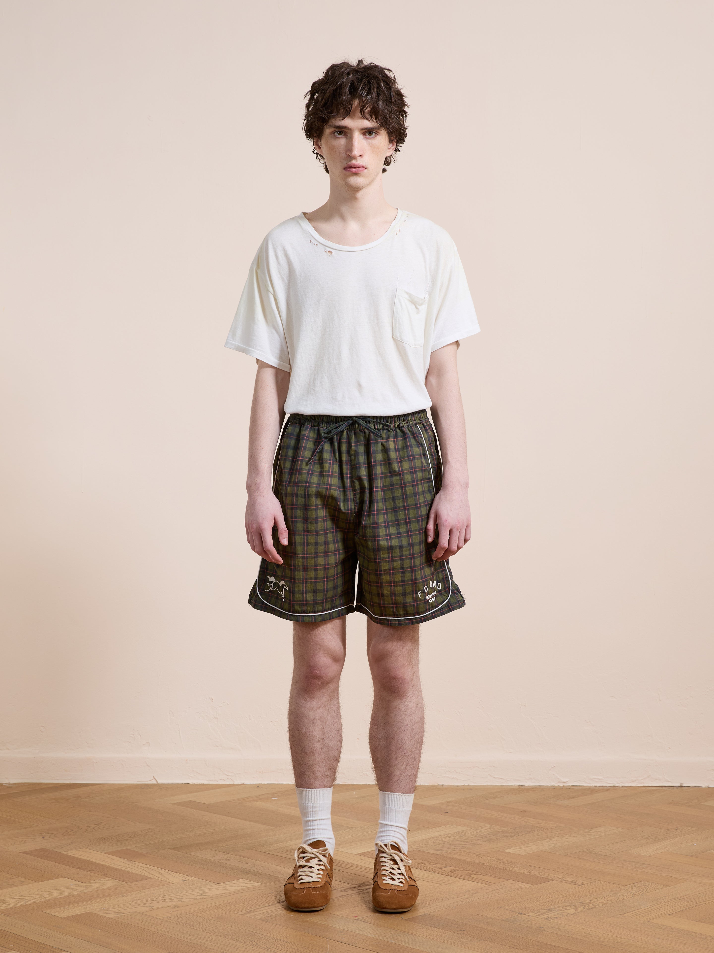 A young person stands indoors on a wooden floor, wearing Found's Forest Plaid Track Shorts with a white t-shirt, white socks, and brown shoes, facing the camera against a plain beige wall.
