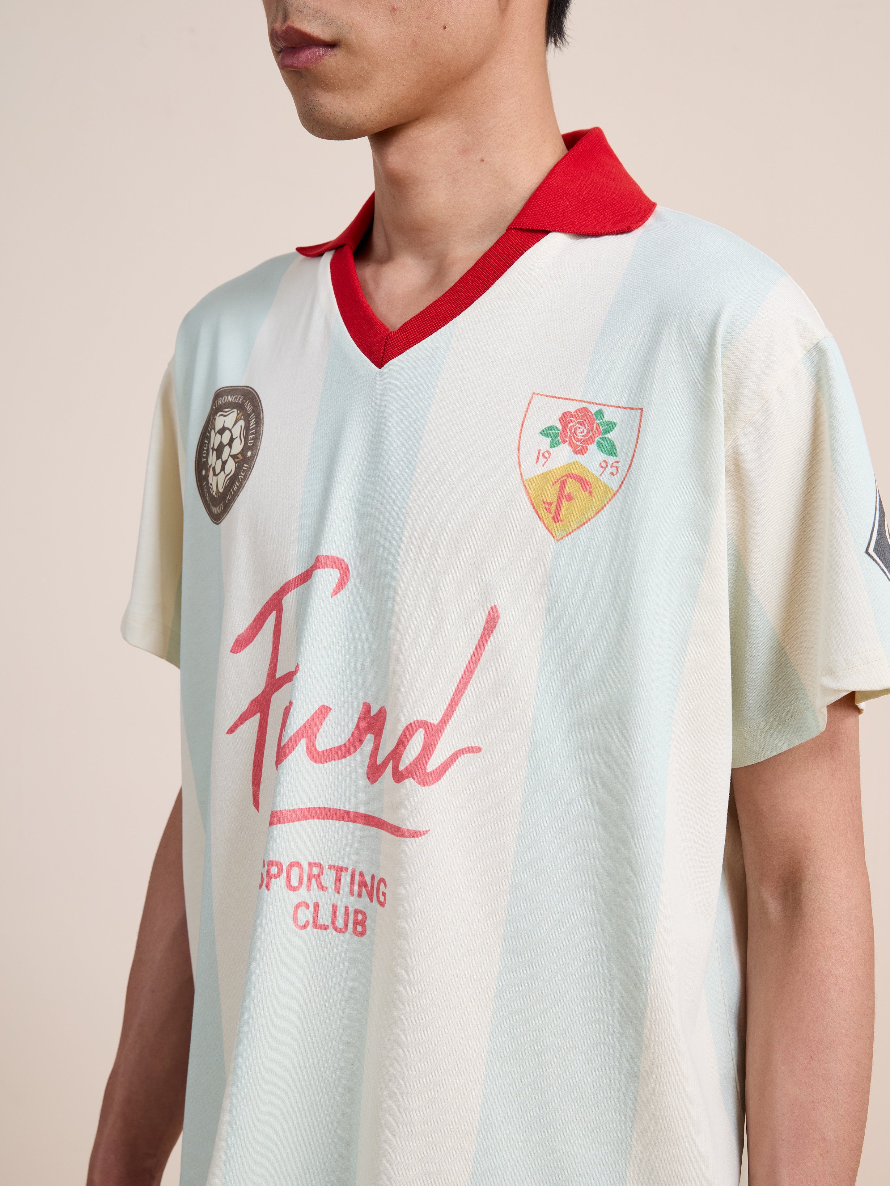 A person wearing the Found Club Short Sleeve Jersey by FOUND—a unisex sportswear piece featuring light blue and white stripes, a red collar, signature logos, and “Fund Sporting Club” text on the front. This jersey captures the essence of a vintage football shirt.