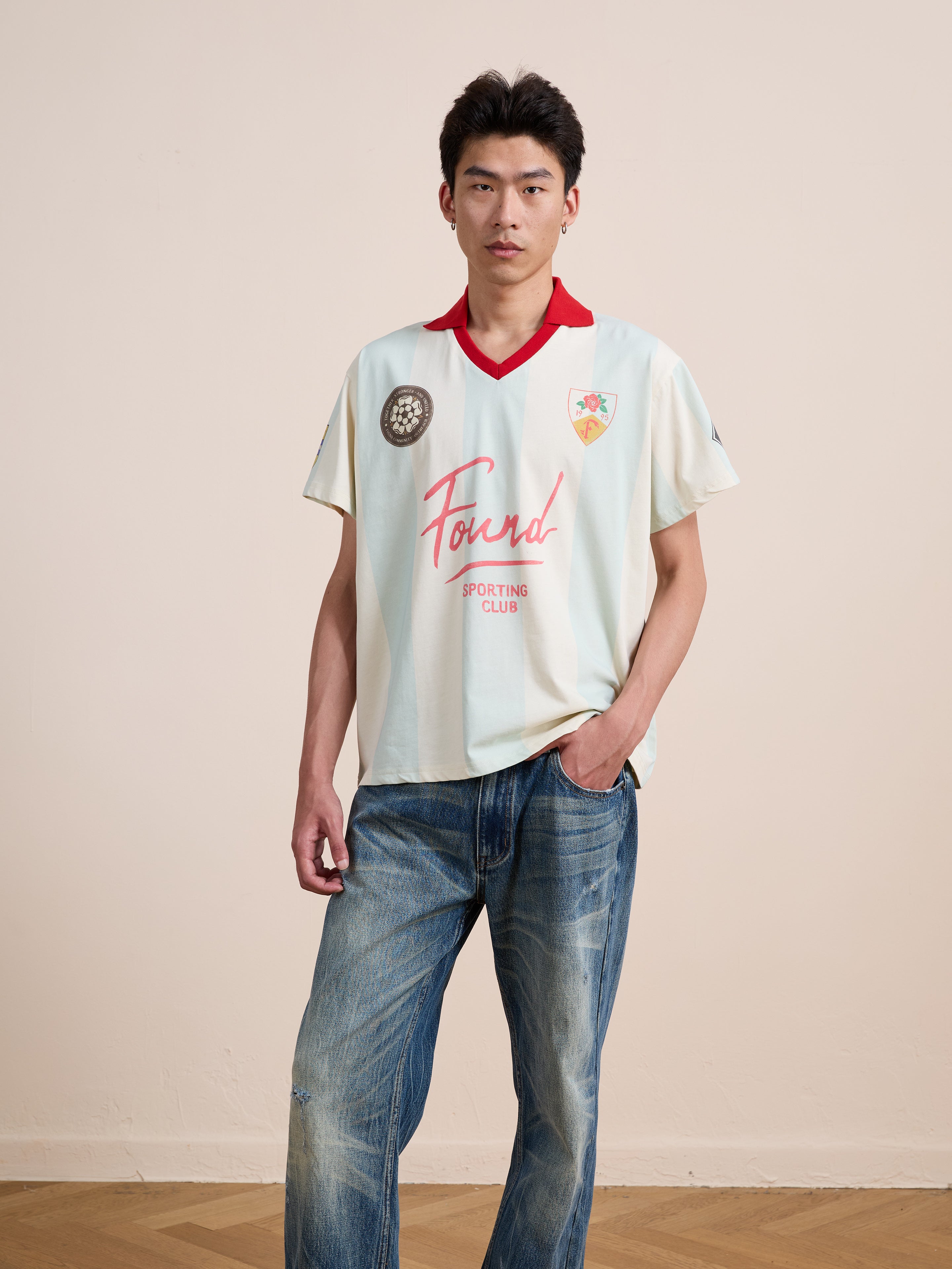 A person stands indoors against a plain wall, wearing the FOUND Found Club Short Sleeve Jersey paired with blue jeans, looking directly at the camera.