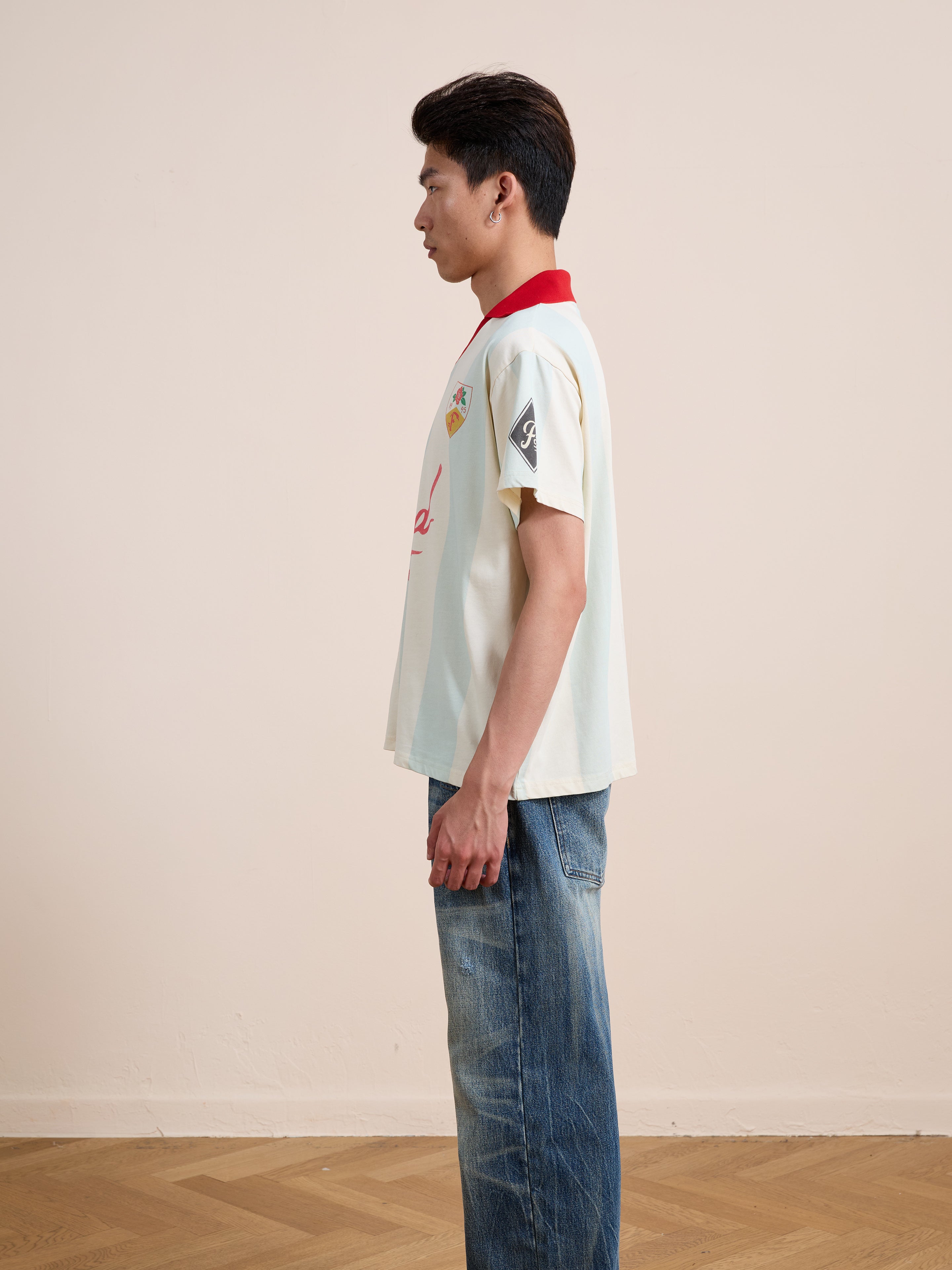 A person stands in profile wearing the FOUND Found Club Short Sleeve Jersey paired with blue jeans in a plain room with a wooden floor.