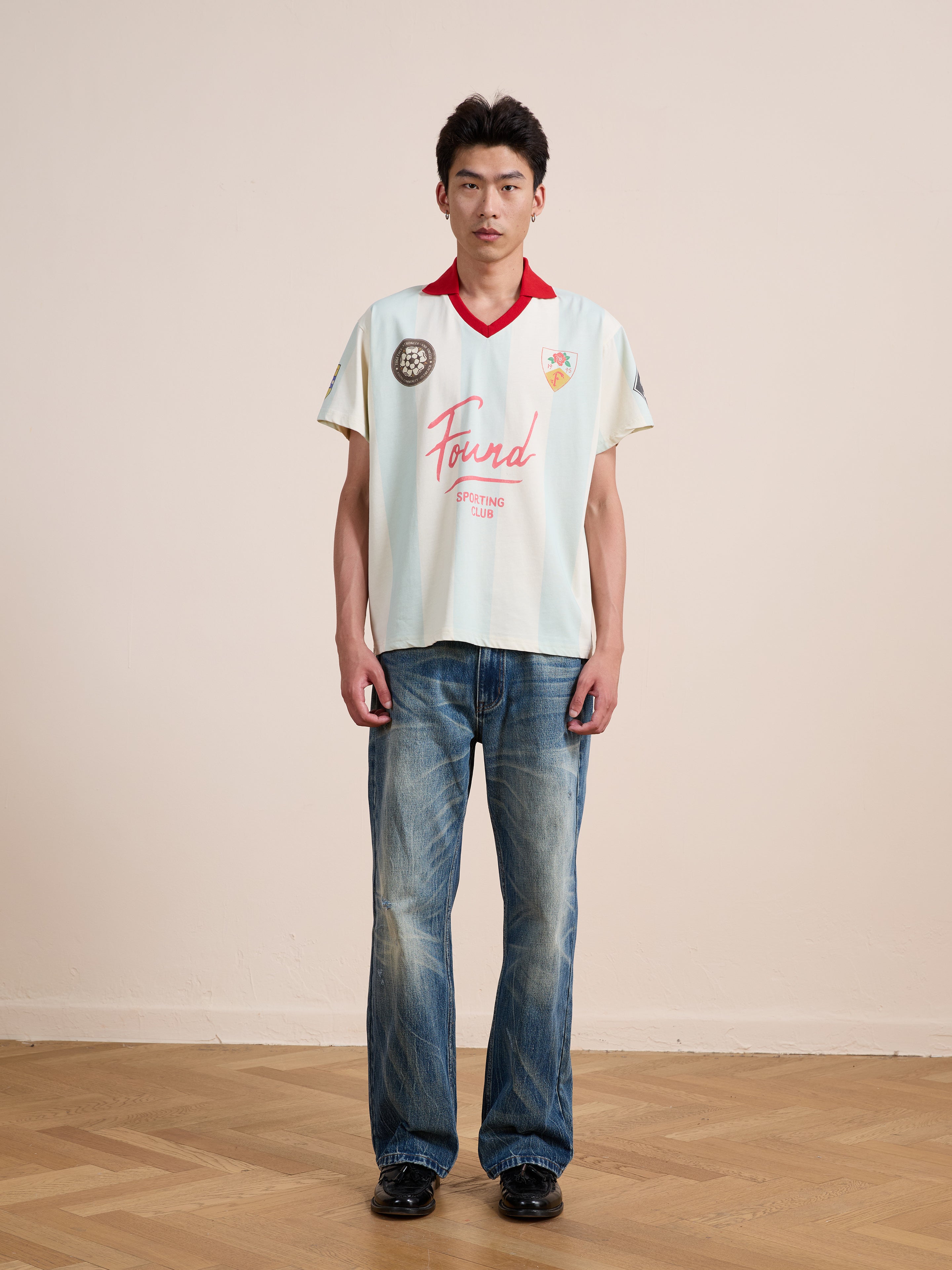 A person stands indoors on a wooden floor with a plain background, wearing the FOUND Found Club Short Sleeve Jersey—a light-colored vintage football shirt with a red collar—paired with blue jeans.
