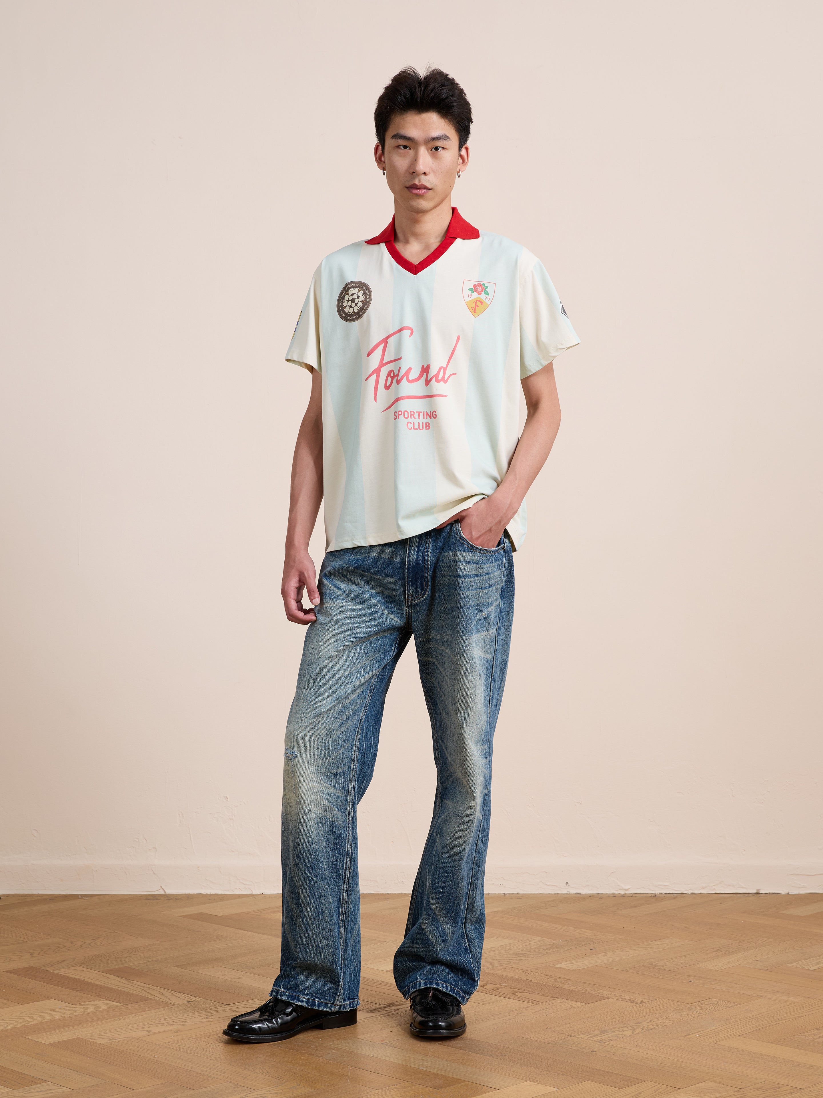 A person stands indoors on a wooden floor against a plain beige wall, wearing a light-colored FOUND Found Club Short Sleeve Jersey paired with blue jeans and black shoes.