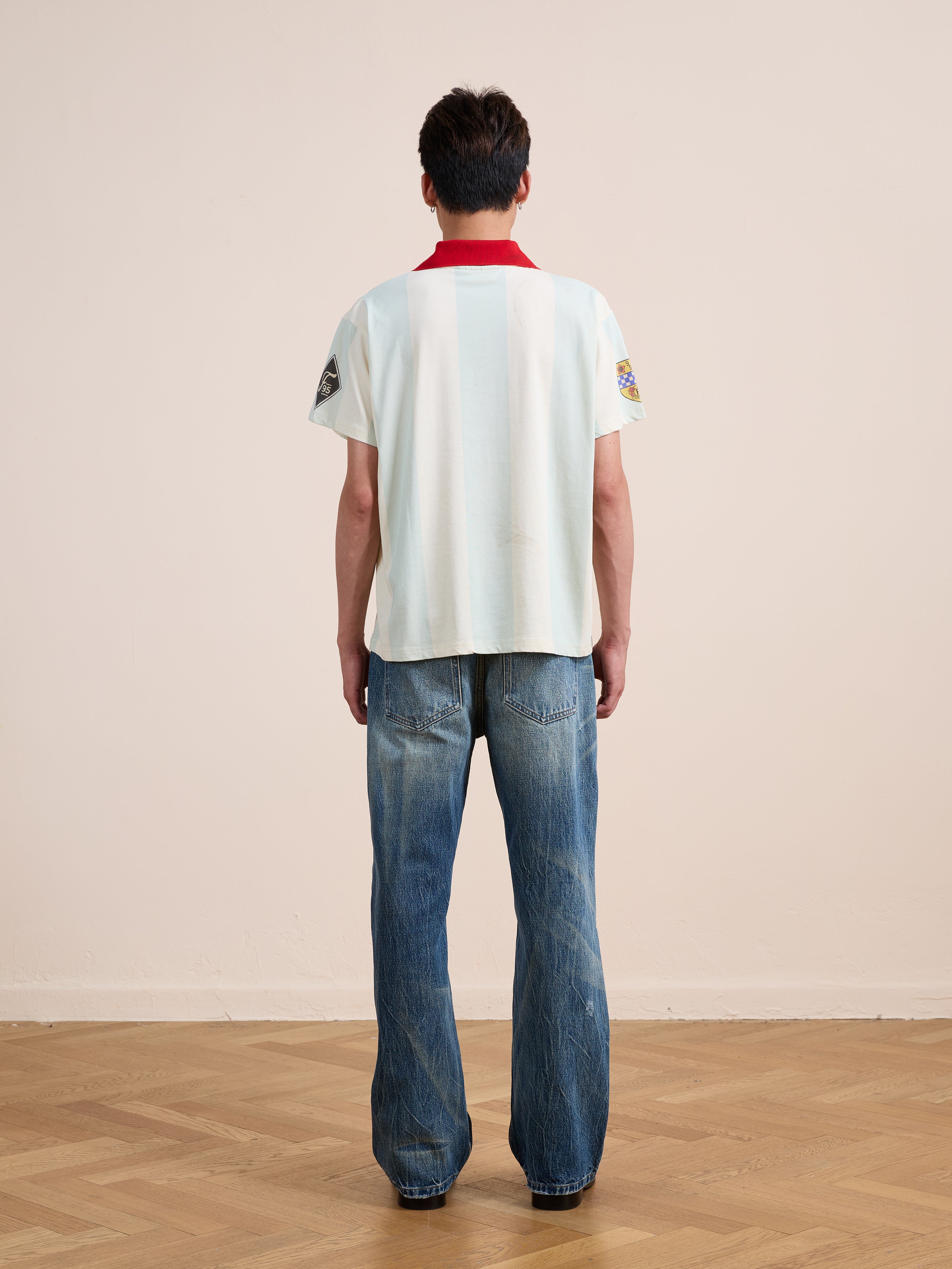 A person stands indoors on a wooden floor, facing away, wearing the FOUND Found Club Short Sleeve Jersey with a red collar and blue jeans.