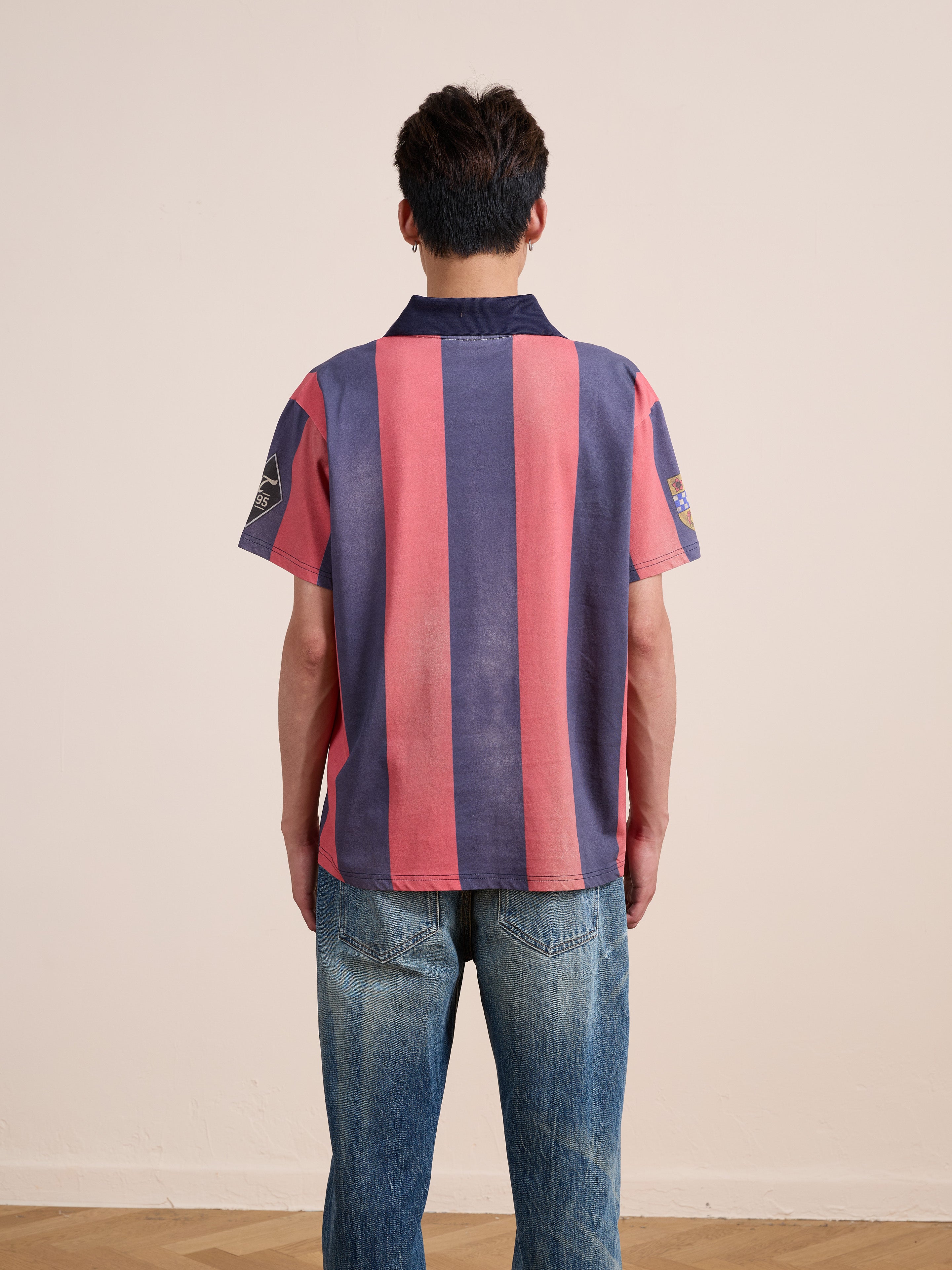 A person stands with their back to the camera, wearing the FOUND Found Club Short Sleeve Jersey—a unisex, relaxed fit top featuring bold red and blue vertical stripes—paired with blue jeans in a simple indoor setting.