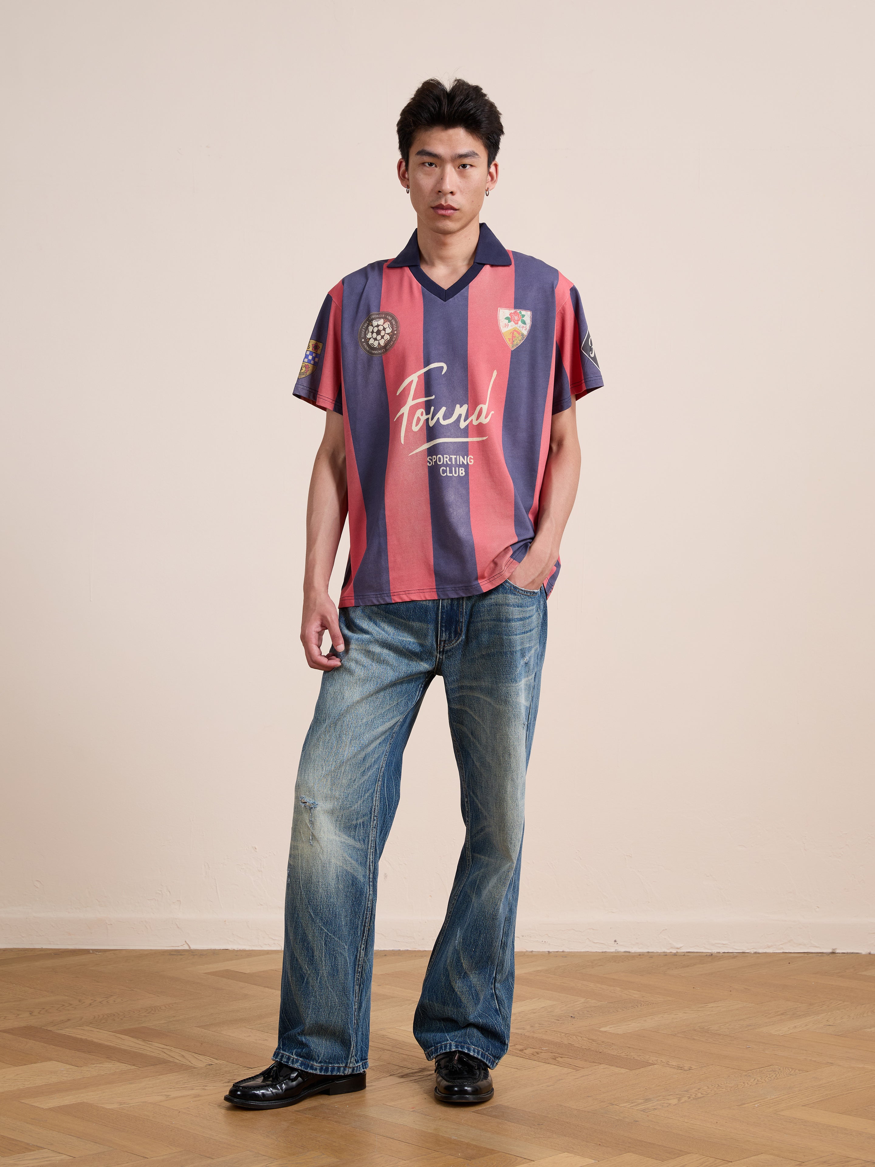 A person stands indoors wearing the FOUND Found Club Short Sleeve Jersey, featuring red and blue stripes in a vintage football shirt style, paired with blue jeans and black shoes on a wooden floor with a plain light-colored wall in the background.