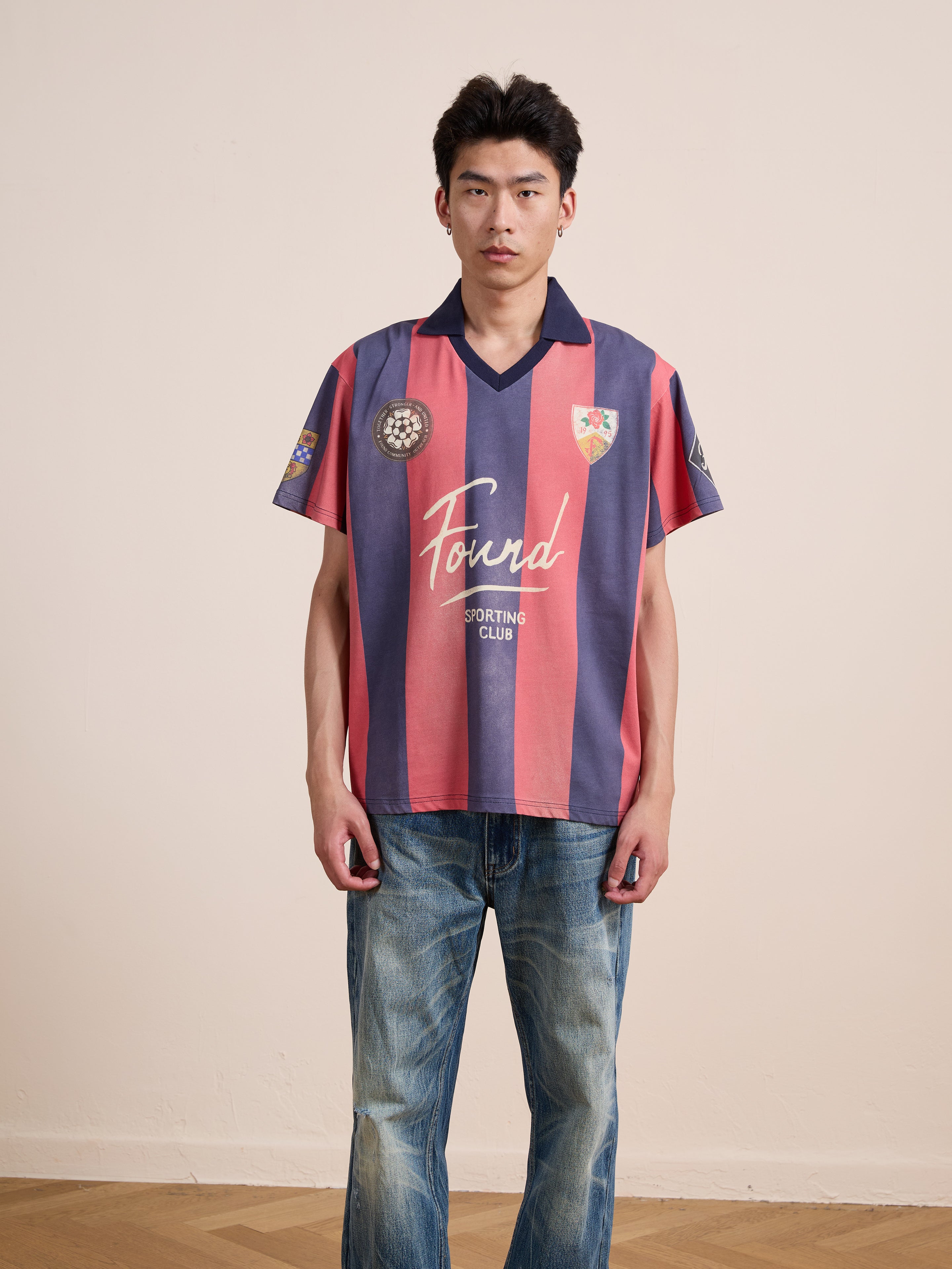 A person stands against a plain wall wearing the FOUND Found Club Short Sleeve Jersey—an unisex, relaxed fit top with red and navy stripes and “Found Sporting Club” text—paired with blue jeans.