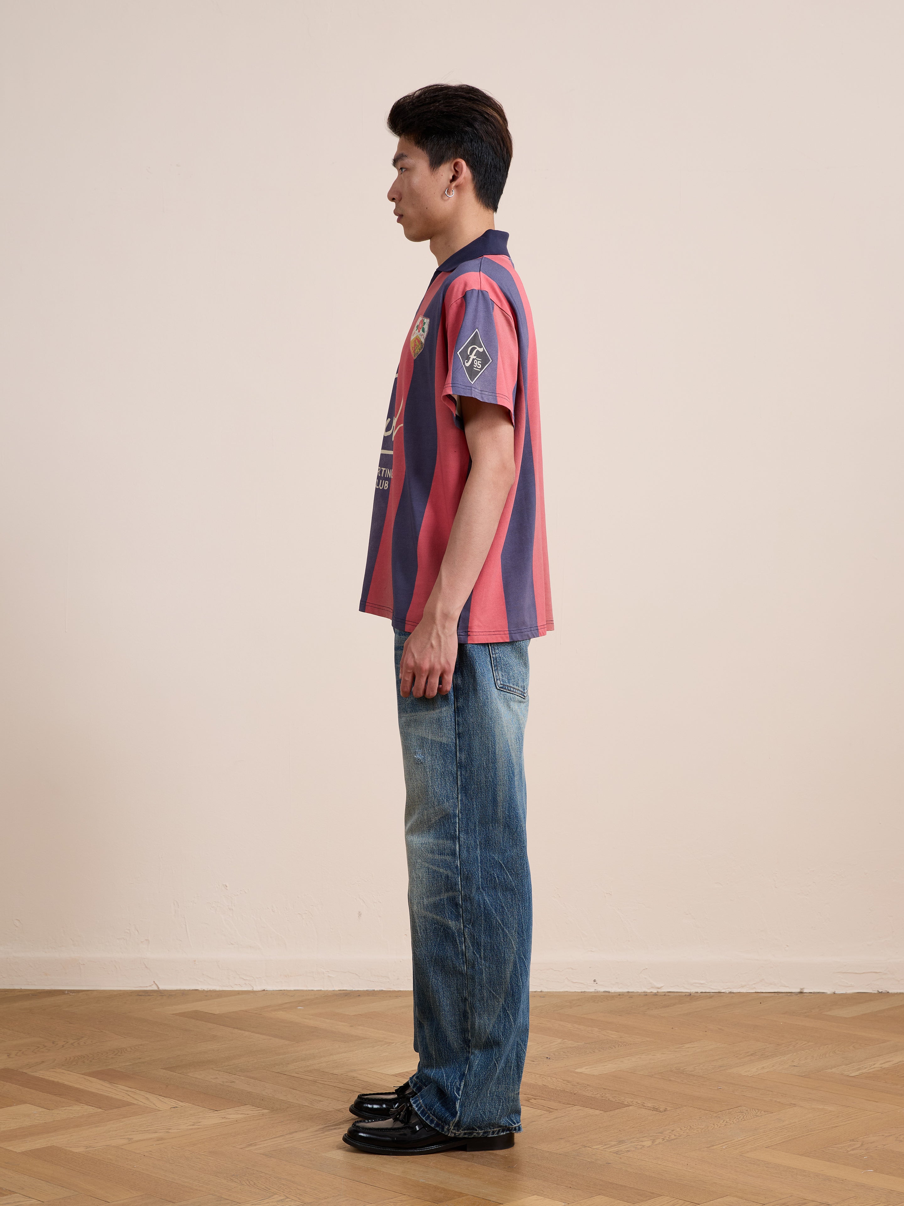 A person stands in profile against a plain light-colored wall and wooden floor, wearing the FOUND Found Club Short Sleeve Jersey styled in red and blue stripes, paired with blue jeans and black shoes.
