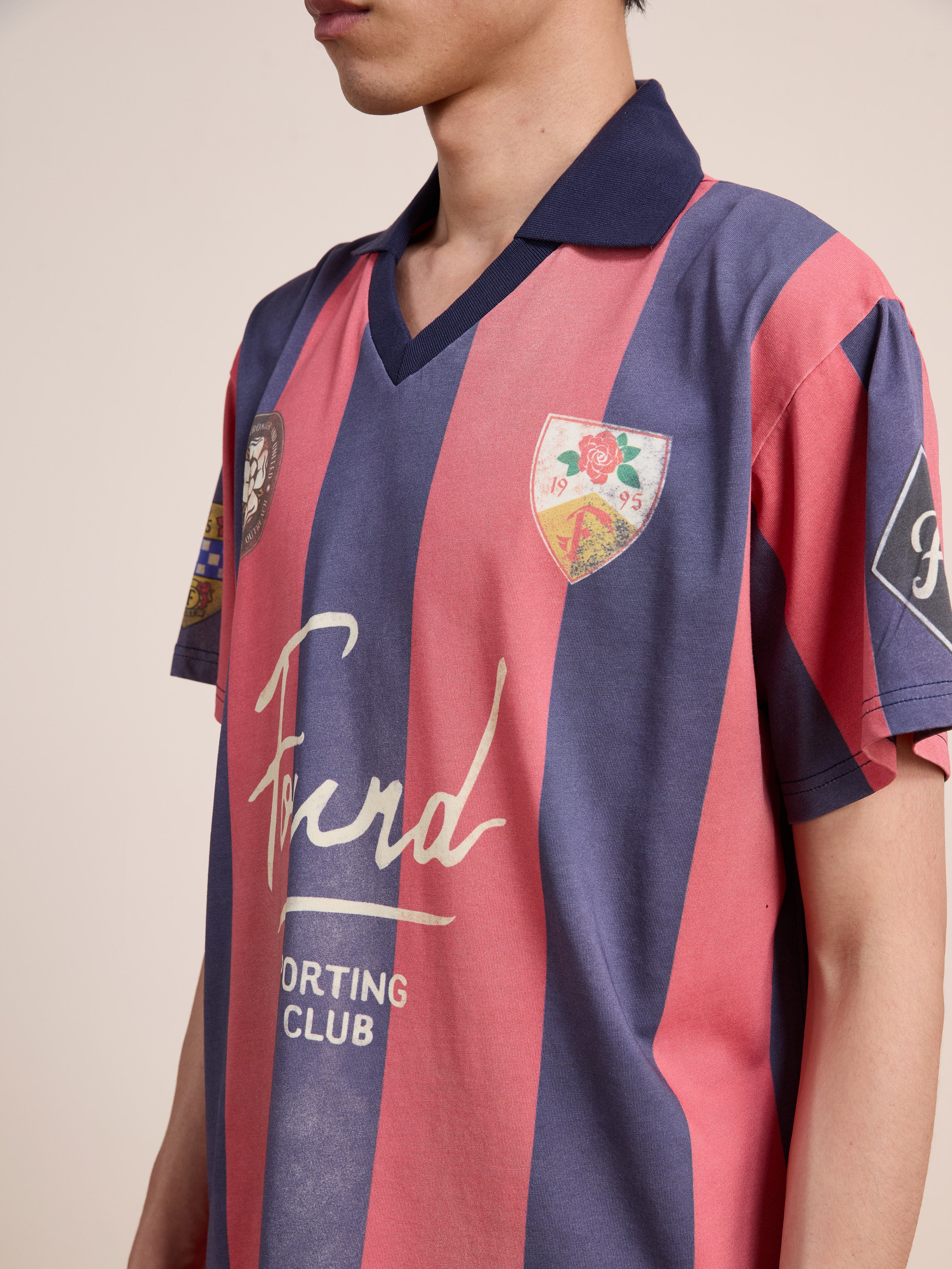 A person wearing the FOUND Found Club Short Sleeve Jersey—a unisex, relaxed fit vintage football shirt in blue and red stripes, featuring badges and “Ford Sporting Club” text on the front.