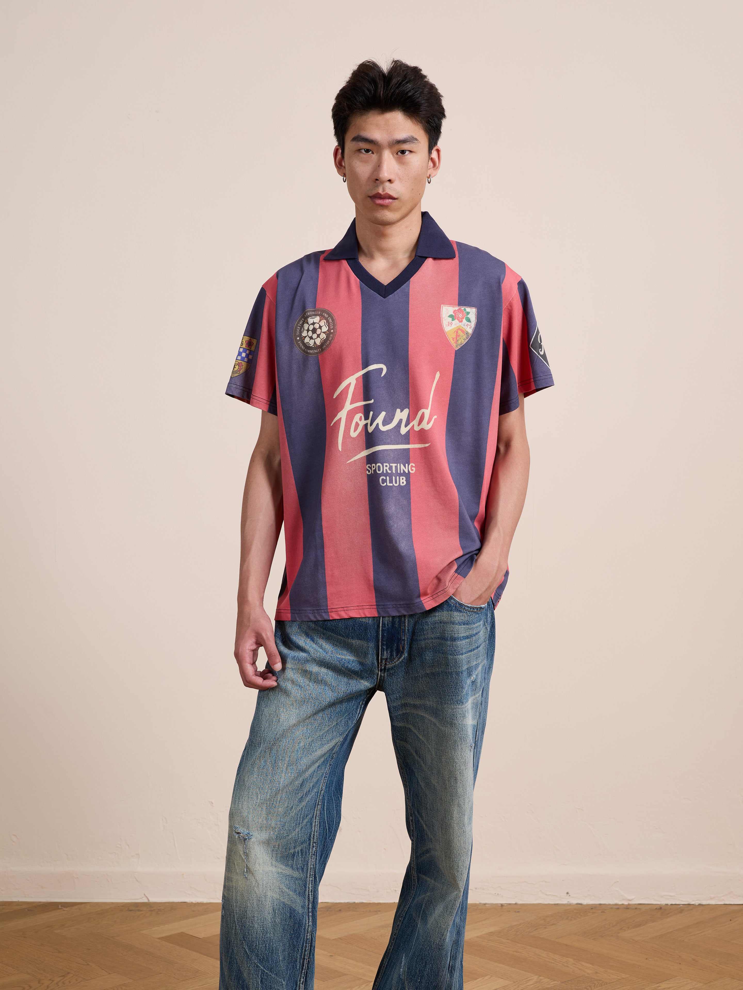 A person stands indoors against a plain background, wearing the FOUND Found Club Short Sleeve Jersey—a unisex vintage football shirt featuring pink and navy stripes—in a relaxed fit, paired with blue jeans and one hand in their pocket.