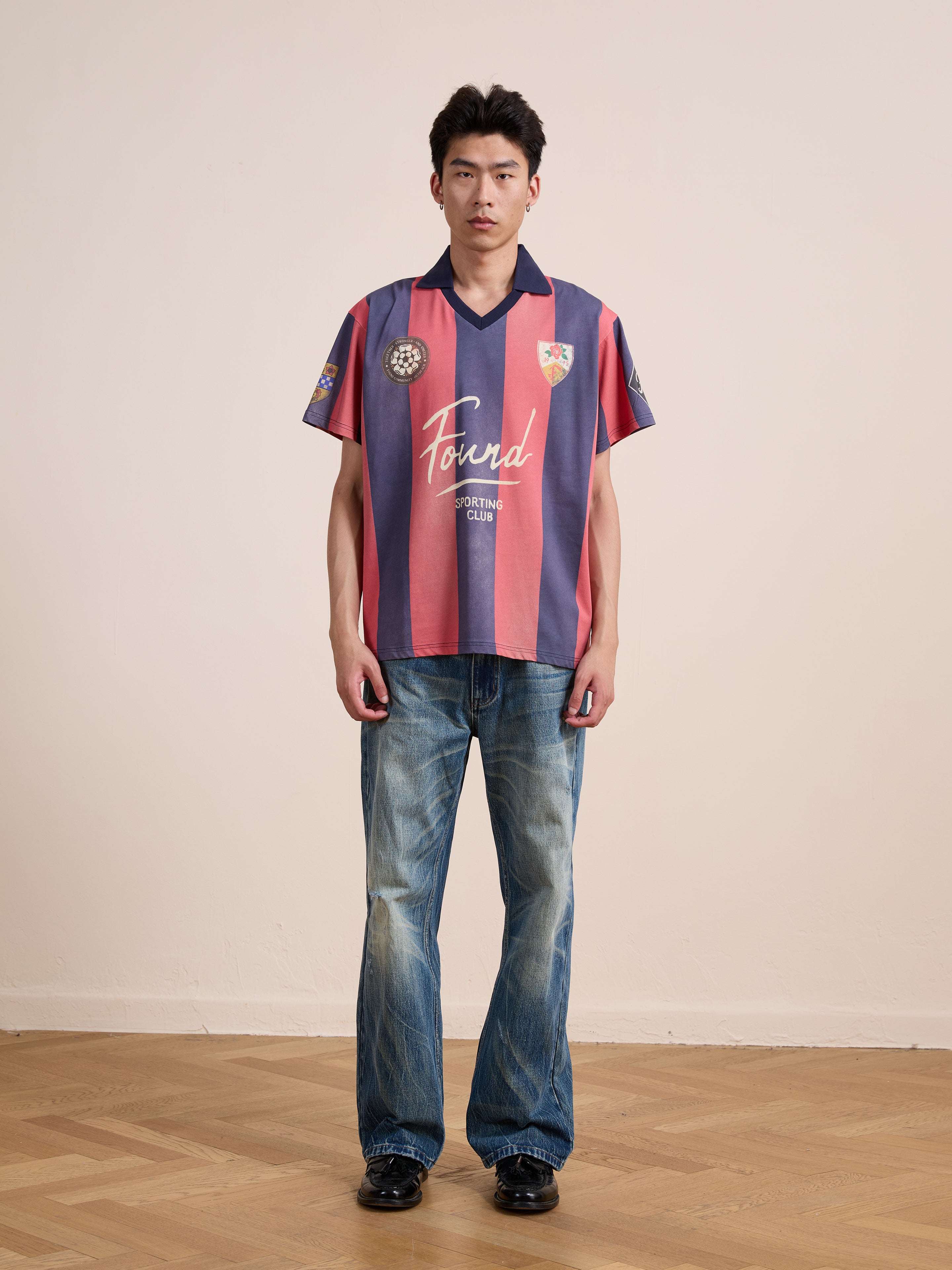 A person stands indoors against a plain beige wall and wooden floor, wearing the FOUND Found Club Short Sleeve Jersey—a red and navy striped unisex relaxed fit top—paired with faded blue jeans and black shoes.
