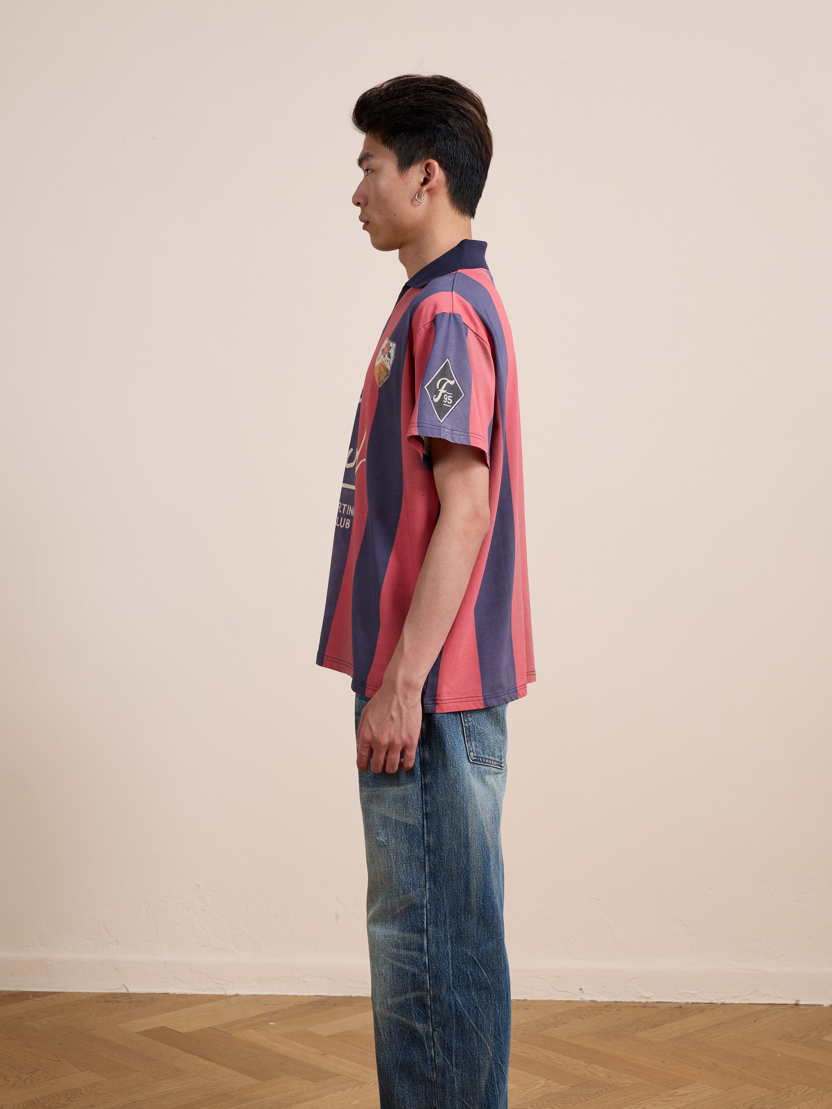 A person stands in profile against a plain beige wall, wearing the FOUND Found Club Short Sleeve Jersey—a unisex relaxed fit striped shirt—paired with blue jeans on a wooden floor.