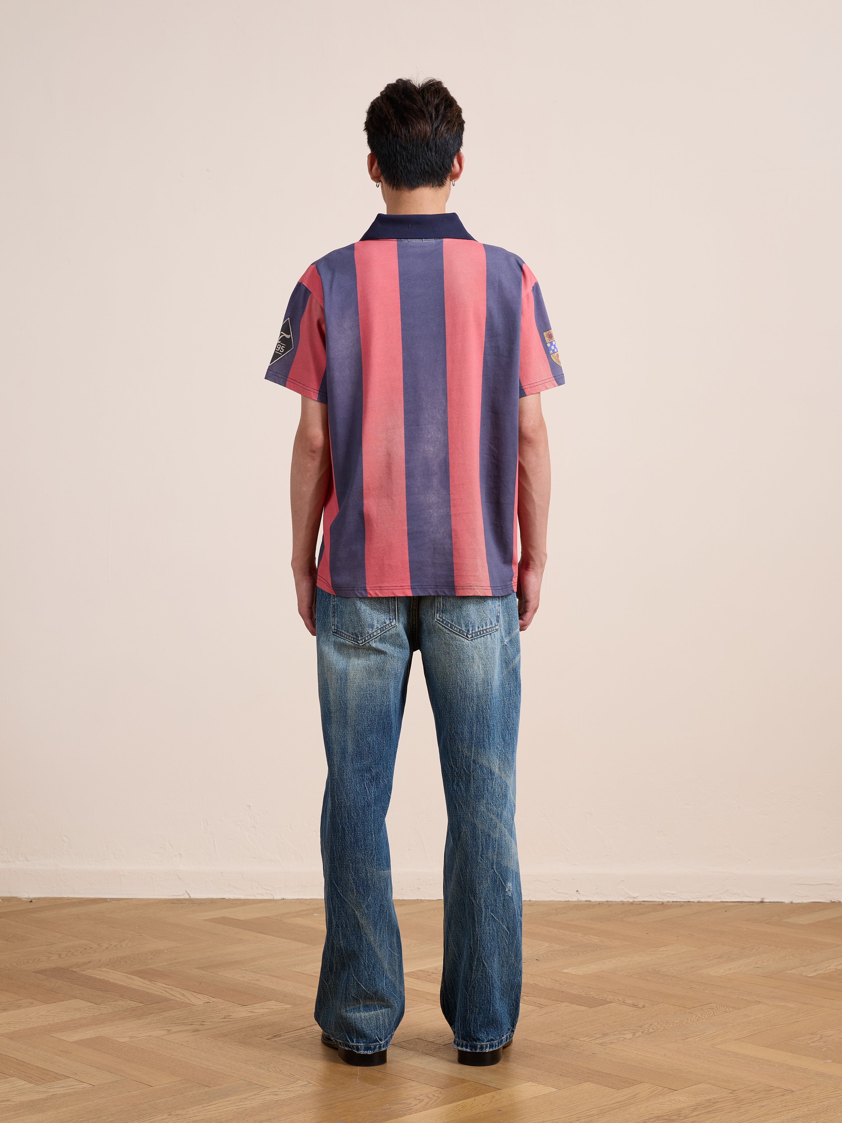A person stands indoors on a wooden floor, facing away, wearing the FOUND Found Club Short Sleeve Jersey—a unisex, relaxed fit shirt with pink and navy vertical stripes reminiscent of a vintage football shirt—and faded blue jeans.