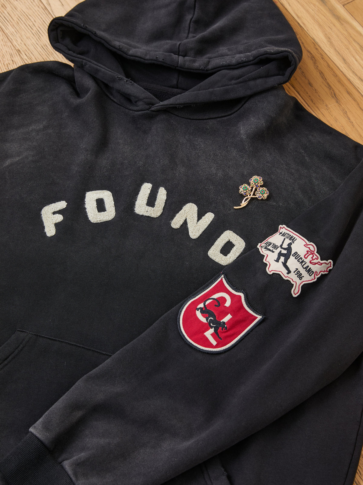The FOUND Found Logo Patch Hoodie, a unisex relaxed fit staple, features black washed cotton fabric with “FOUND” text, a tree-shaped pin, a red horse crest patch, and a “Where Brooklyn at?!” patch on the sleeve. Shown laid flat on a wooden surface, it combines unique details with effortless comfort for everyday wear.
