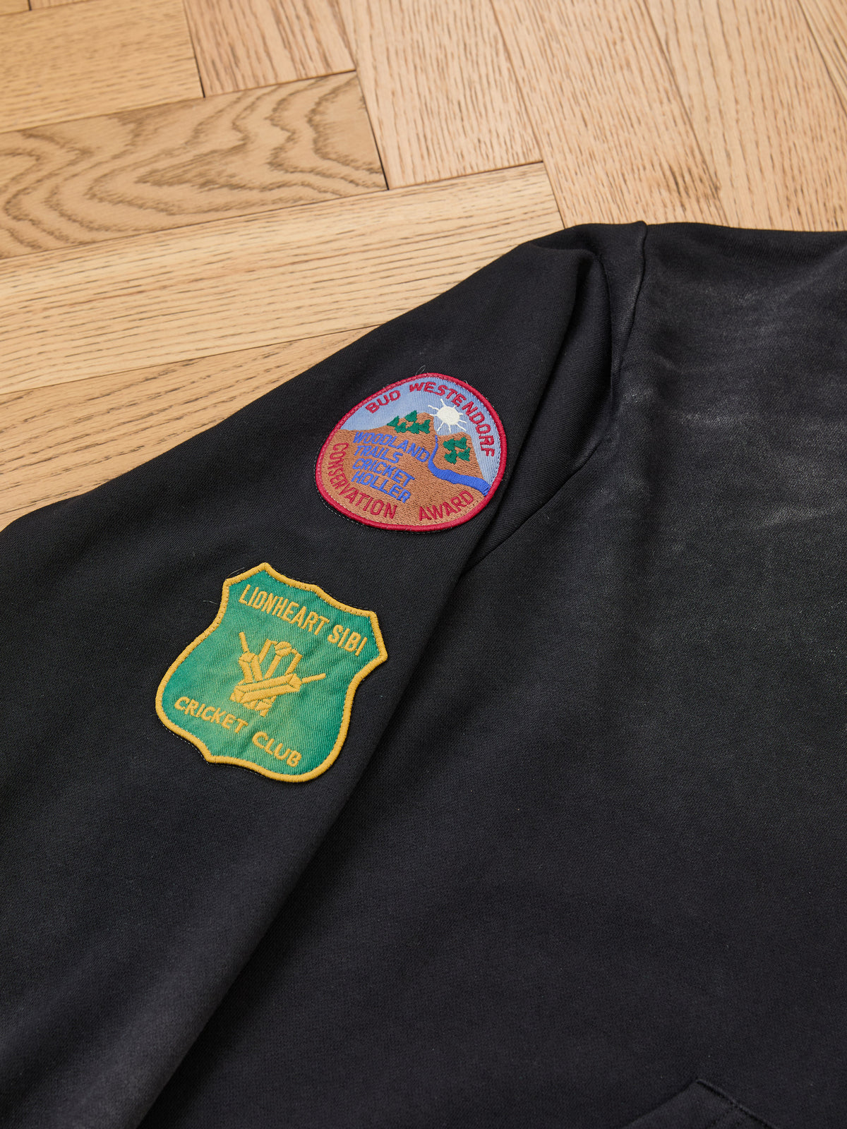 A black sleeve from the FOUND Found Logo Patch Hoodie, a unisex relaxed fit style, displays two patches: a round patch with a red border and a green shield-shaped patch labeled “Lionheart Sibu Cricket Club,” shown against a wooden floor background.