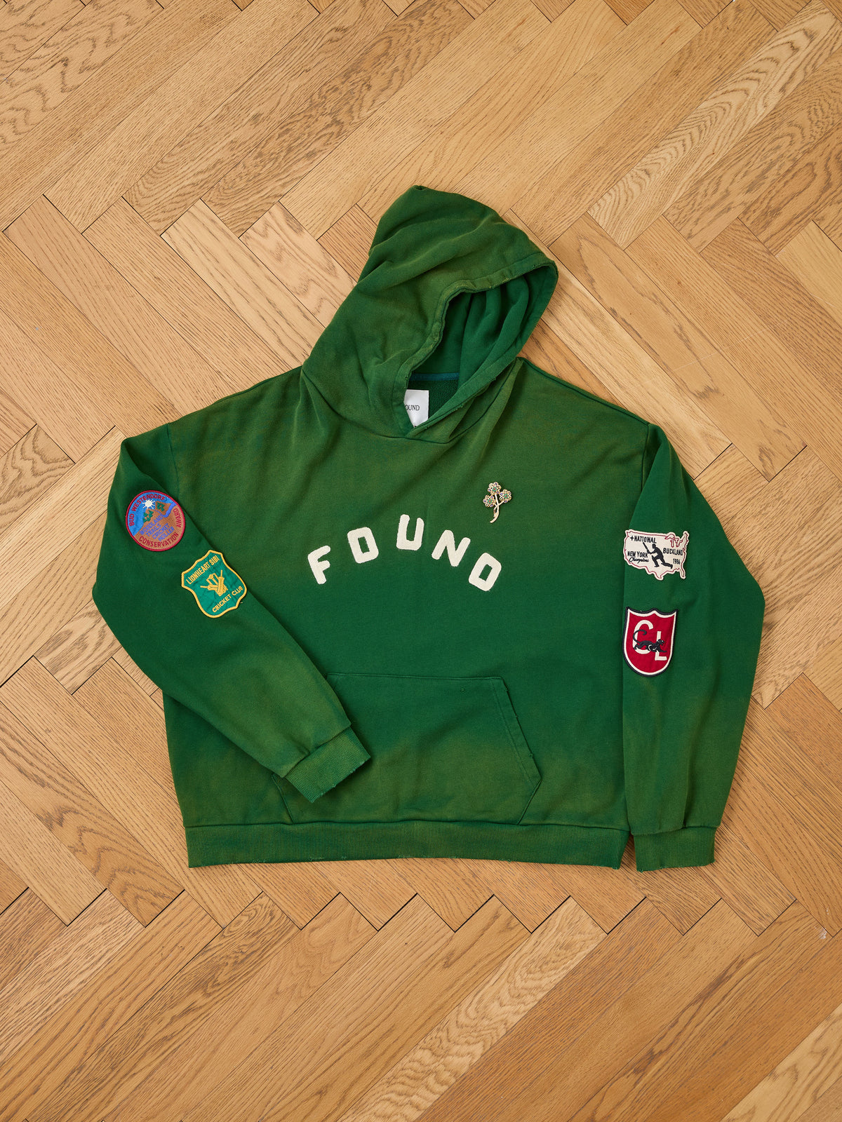 FOUND’s Found Logo Patch Hoodie in vintage forest green, unisex relaxed fit, features “FOUND” text and colorful logo patches. Shown laid flat on a wooden floor.