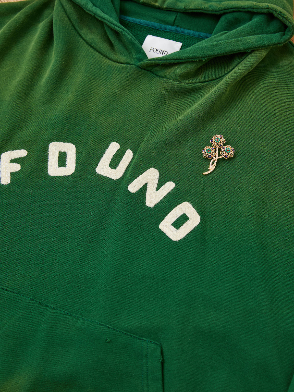 The Found Logo Patch Hoodie by FOUND is a vintage forest green unisex relaxed fit hoodie with white “FOUND” lettering and a clover-shaped chest pin.