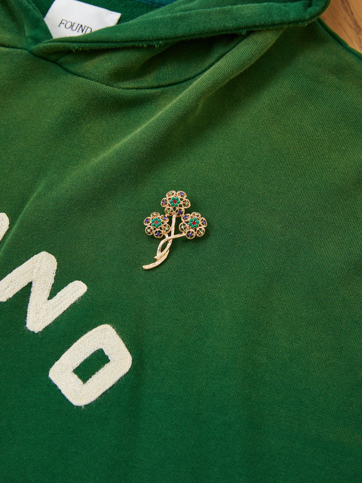 A gold and jeweled tree-shaped brooch is pinned to a FOUND Found Logo Patch Hoodie in vintage forest green with partially visible white text.
