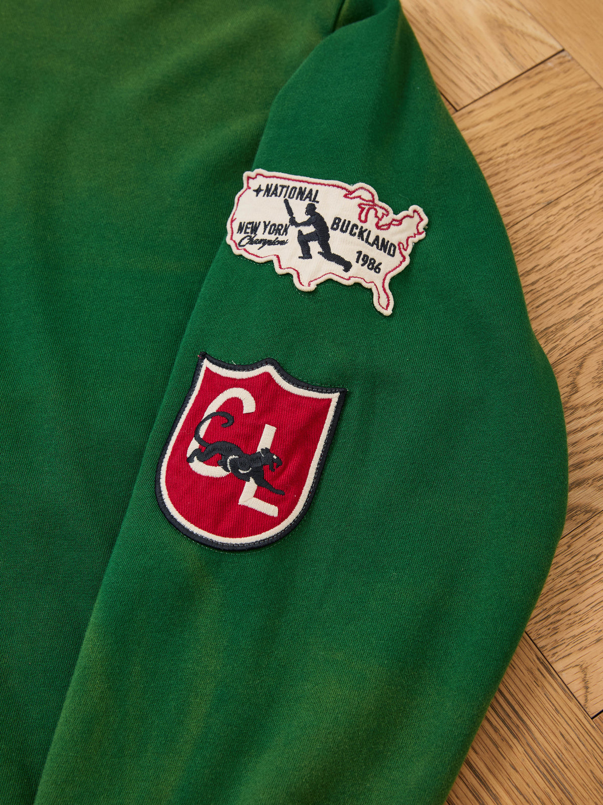 The Found Logo Patch Hoodie by FOUND, in vintage forest green with two distinctive patches, rests on a wooden floor.
