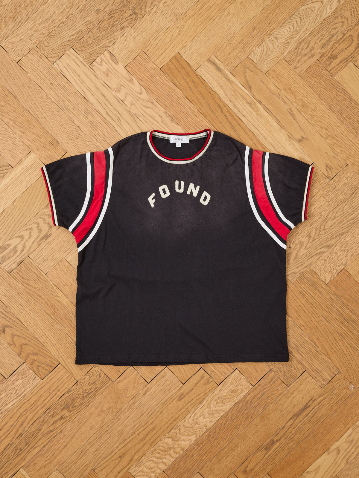 FOUND Foundation Practice Jersey—a black t-shirt with red and white striped sleeves and “FOUND” printed in bold white letters on the front, channeling a retro sportswear vibe—laid flat on a wooden floor.