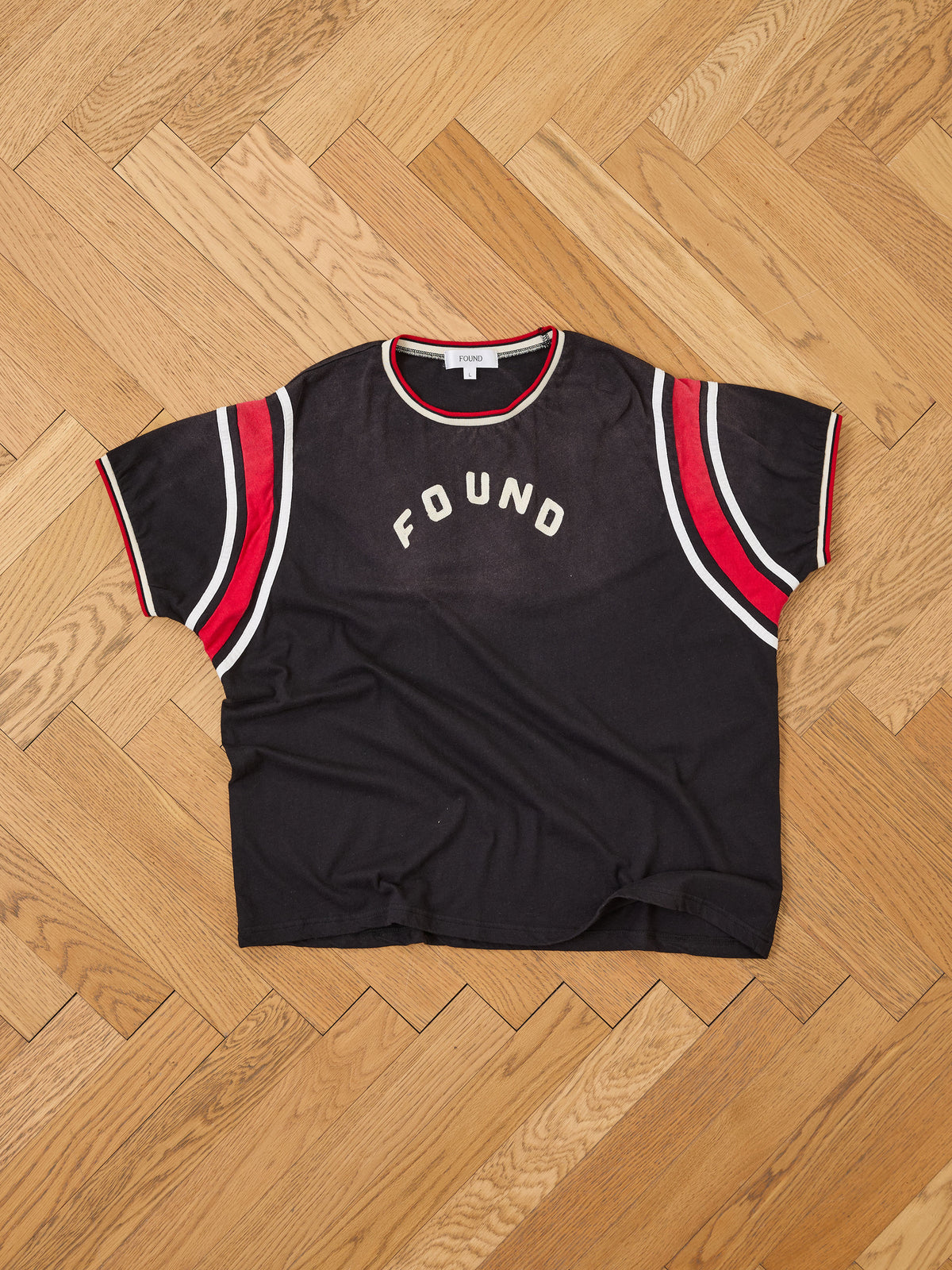 FOUND Foundation Practice Jersey—black T-shirt featuring red and white striped sleeves and “FOUND” printed on the chest, styled like a vintage athletic jersey and displayed flat on a wooden herringbone floor.