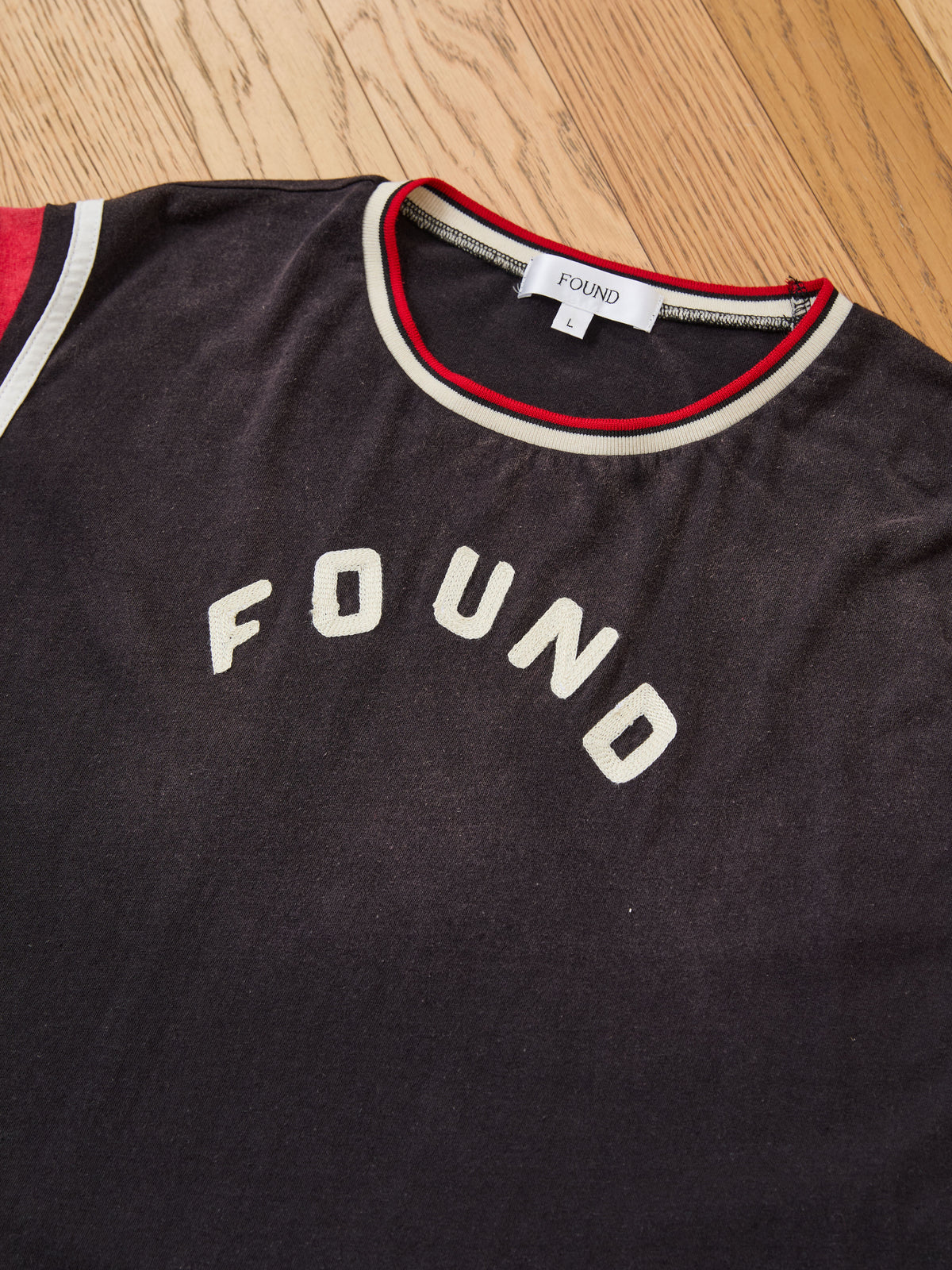 A black Foundation Practice Jersey by FOUND, featuring red and white trim reminiscent of a vintage athletic jersey, displays the word “FOUND” printed on the chest and a visible “FOUND” label at the neck, laid on a wooden floor.
