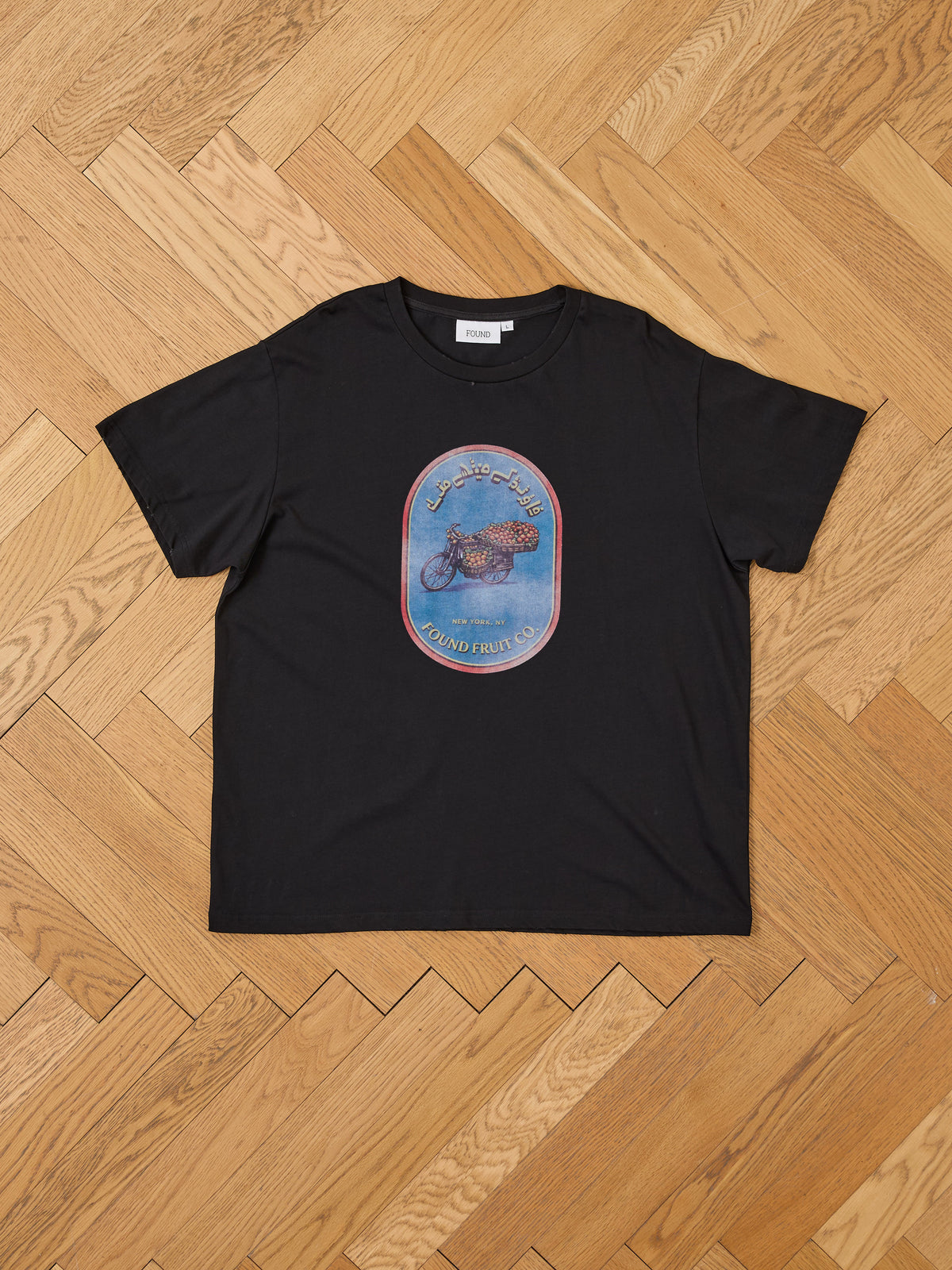 FOUND Fruit Co Tee featuring a colorful fruit bowl and stylized text, unisex relaxed fit, shown laid flat on a light wooden herringbone floor.