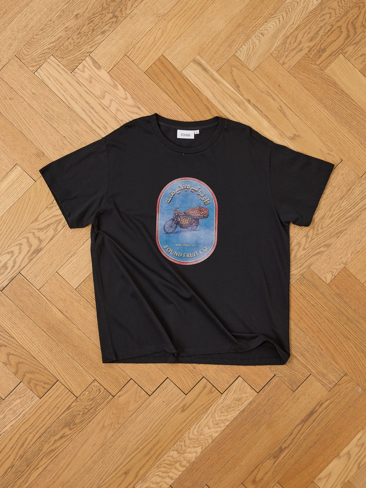 FOUND Fruit Co Tee in black with a vibrant oval graphic on the front, unisex relaxed fit, shown flat on a light wooden herringbone floor.