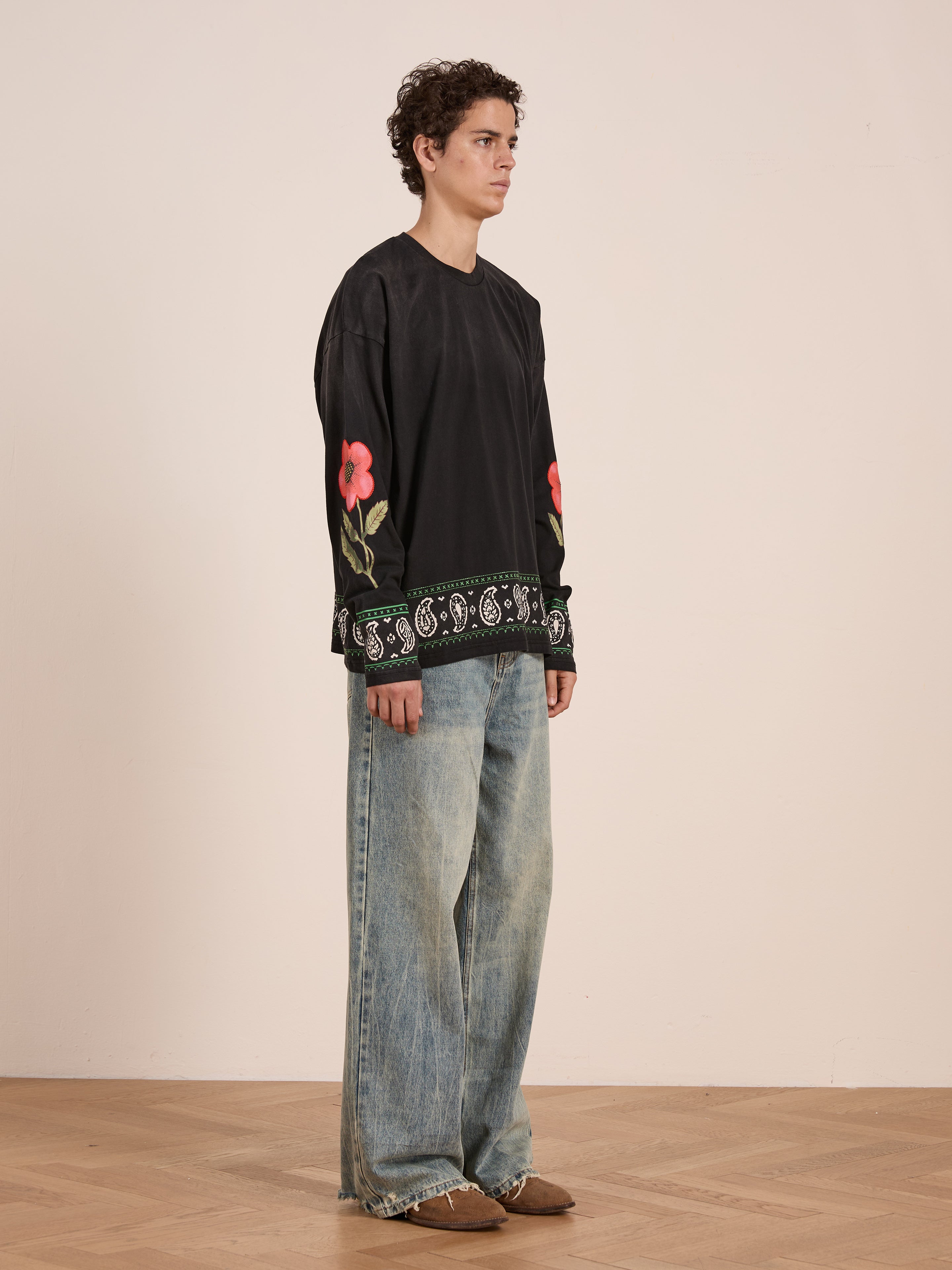 Indoors, a person wears the FOUND Garden Bloom Embroidered LS Tee with floral embroidery on the sleeves and hem, styled with wide-leg faded jeans and brown shoes for a relaxed look.