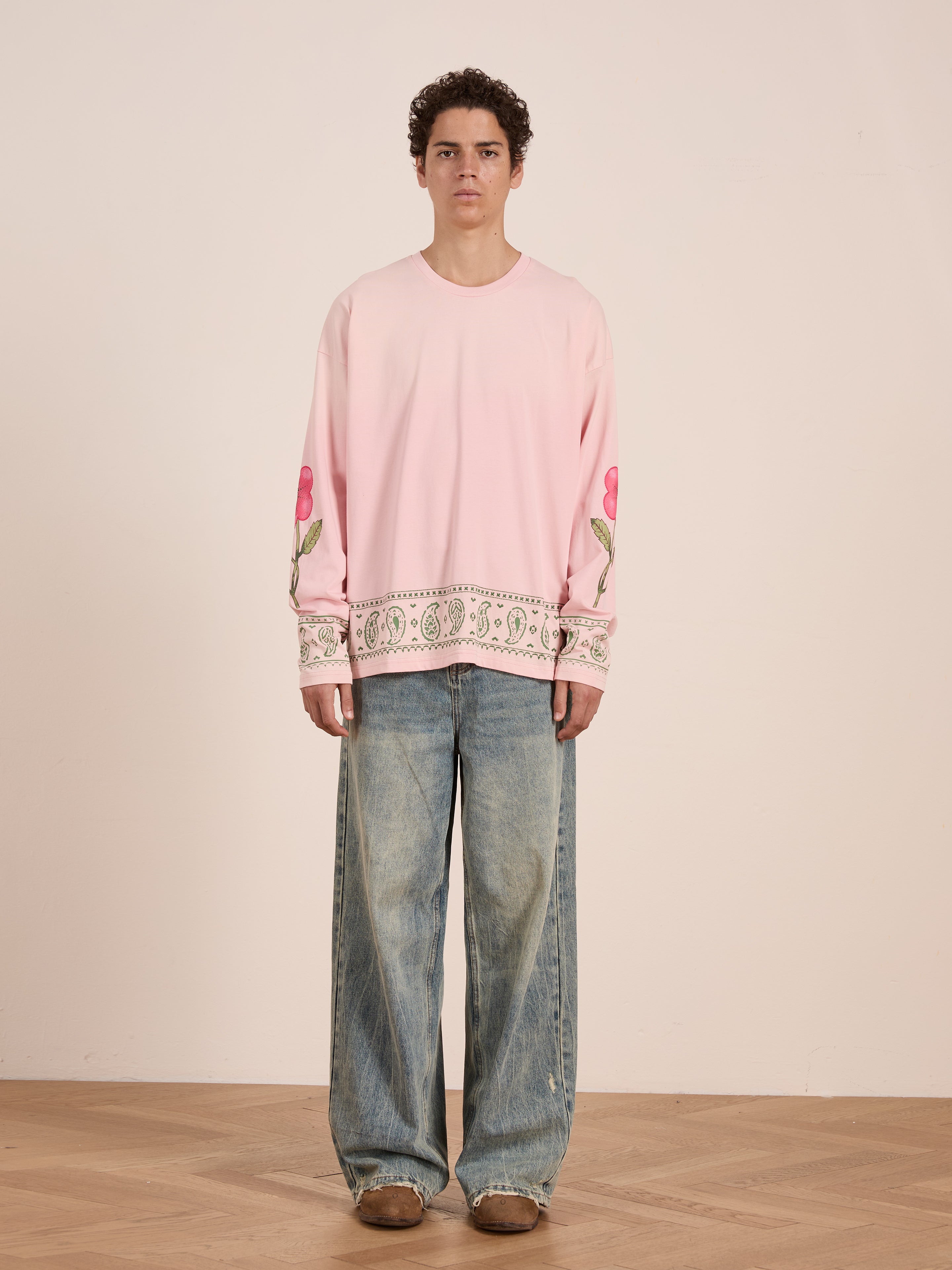 A person stands against a plain wall wearing the FOUND Garden Bloom Embroidered LS Tee with floral sleeve and hem details, unisex relaxed fit wide blue jeans, and brown shoes.