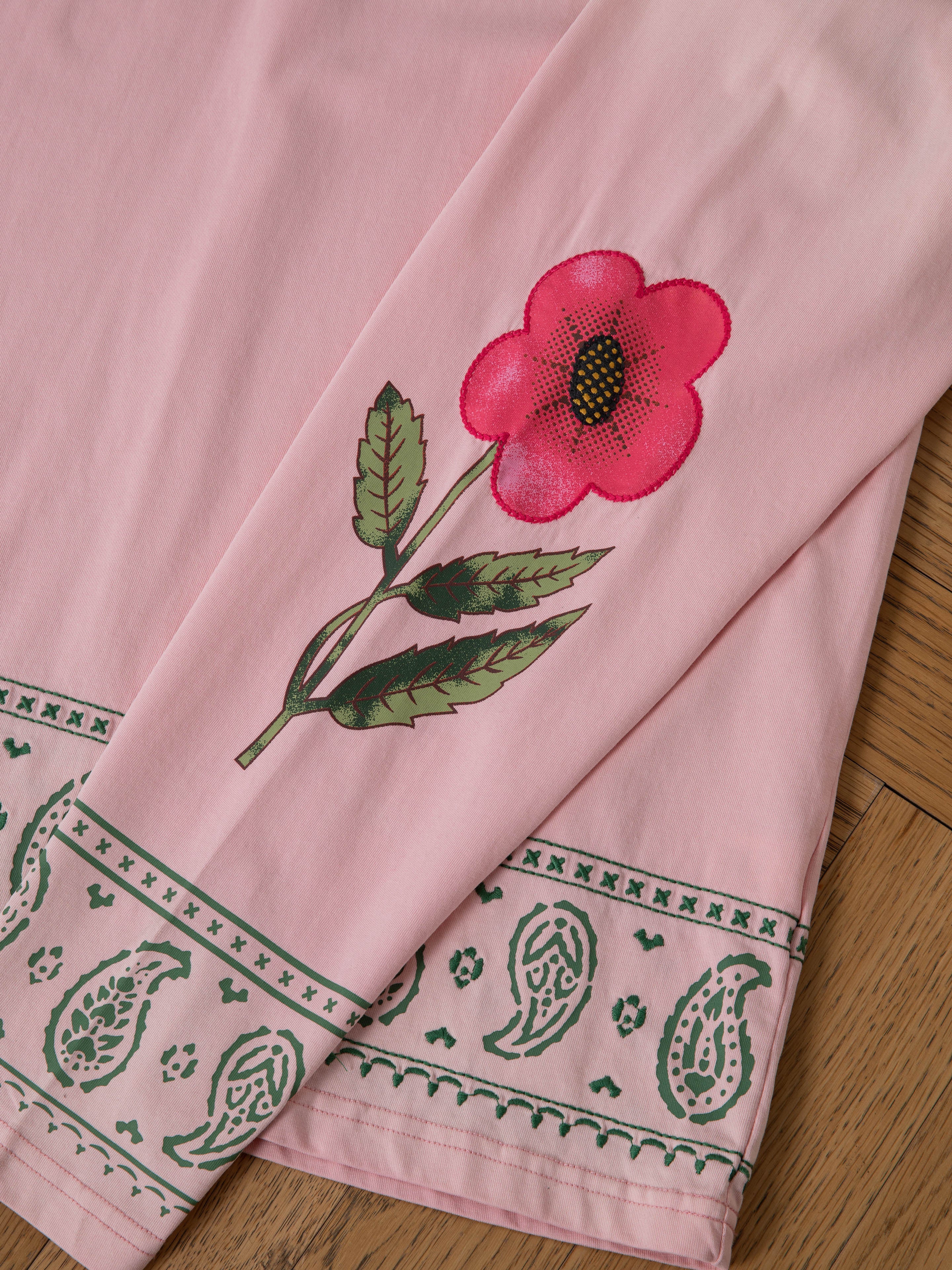 The FOUND Garden Bloom Embroidered LS Tee in pink features floral embroidery with a red flower, green leaves, and a green paisley border on the cuff, displayed on a wooden surface.