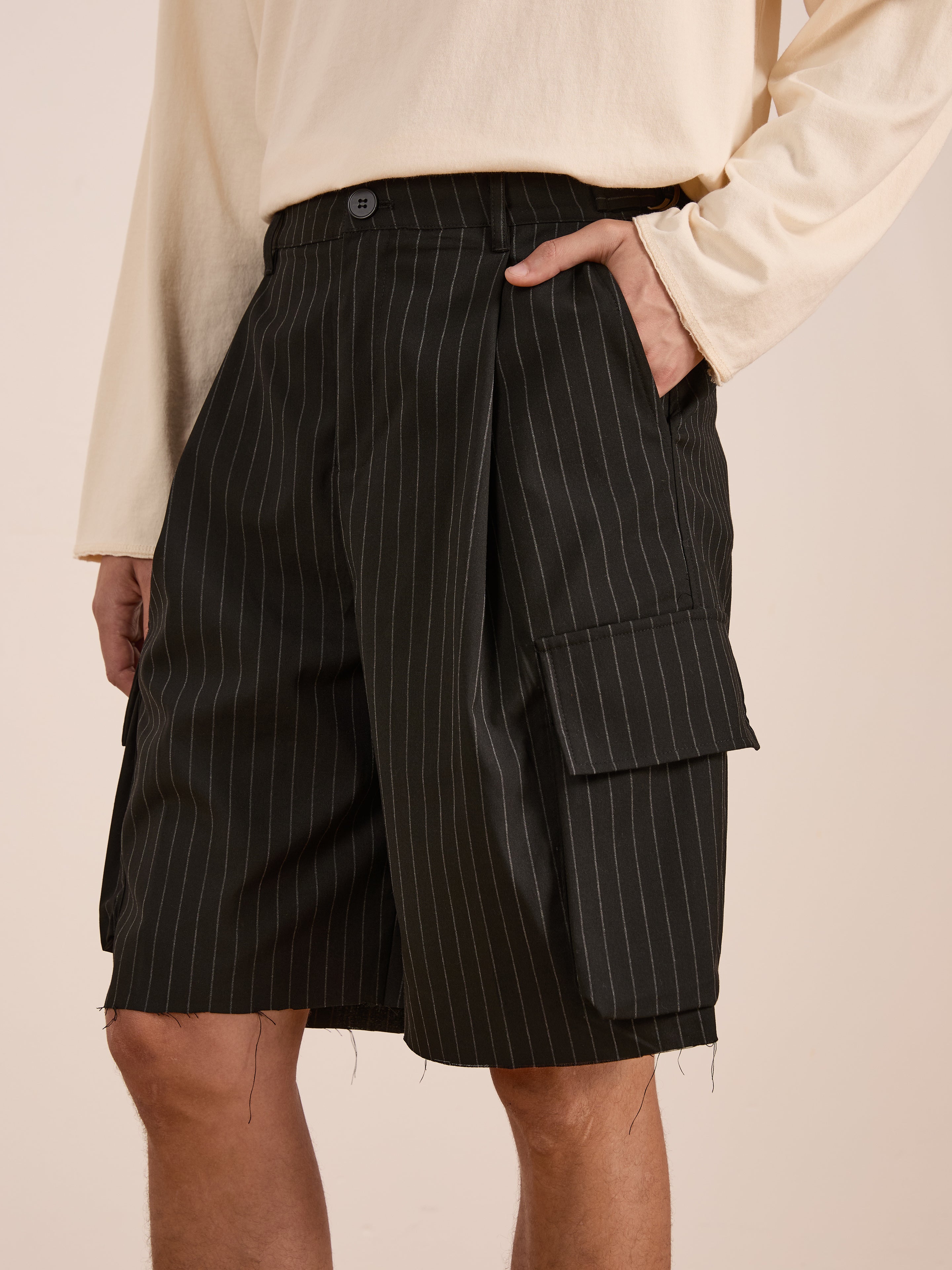 A person wears a cream long-sleeve shirt and FOUND's Hava Pinstripe Pleated Cargo Trouser Shorts, which feature side flap pockets and an adjustable fit for comfort.