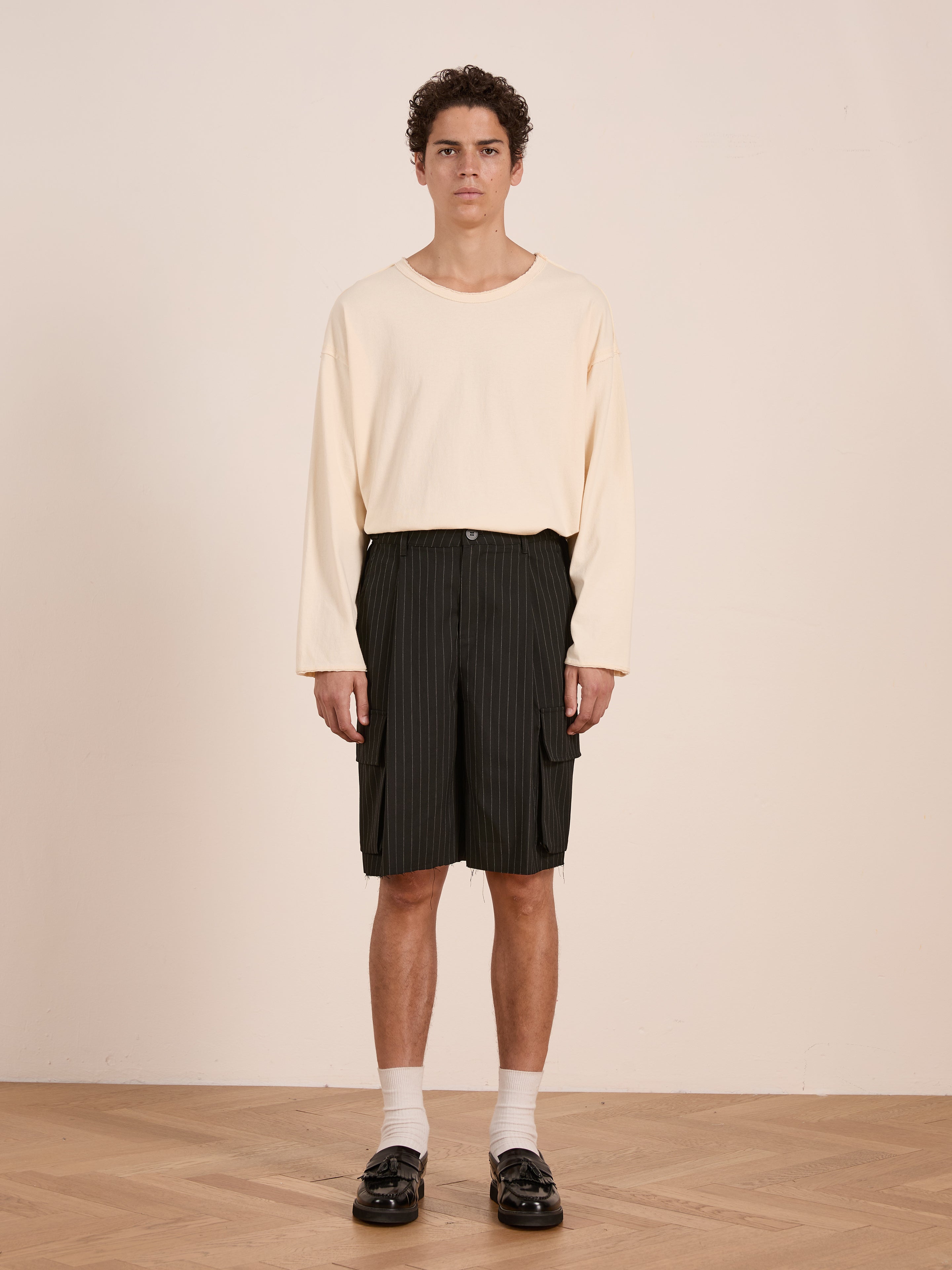 Indoors, a person wears a loose cream long-sleeve shirt, FOUND Hava Pinstripe Pleated Cargo Trouser Shorts, white socks, and black loafers on a wooden floor against a plain light-colored wall.