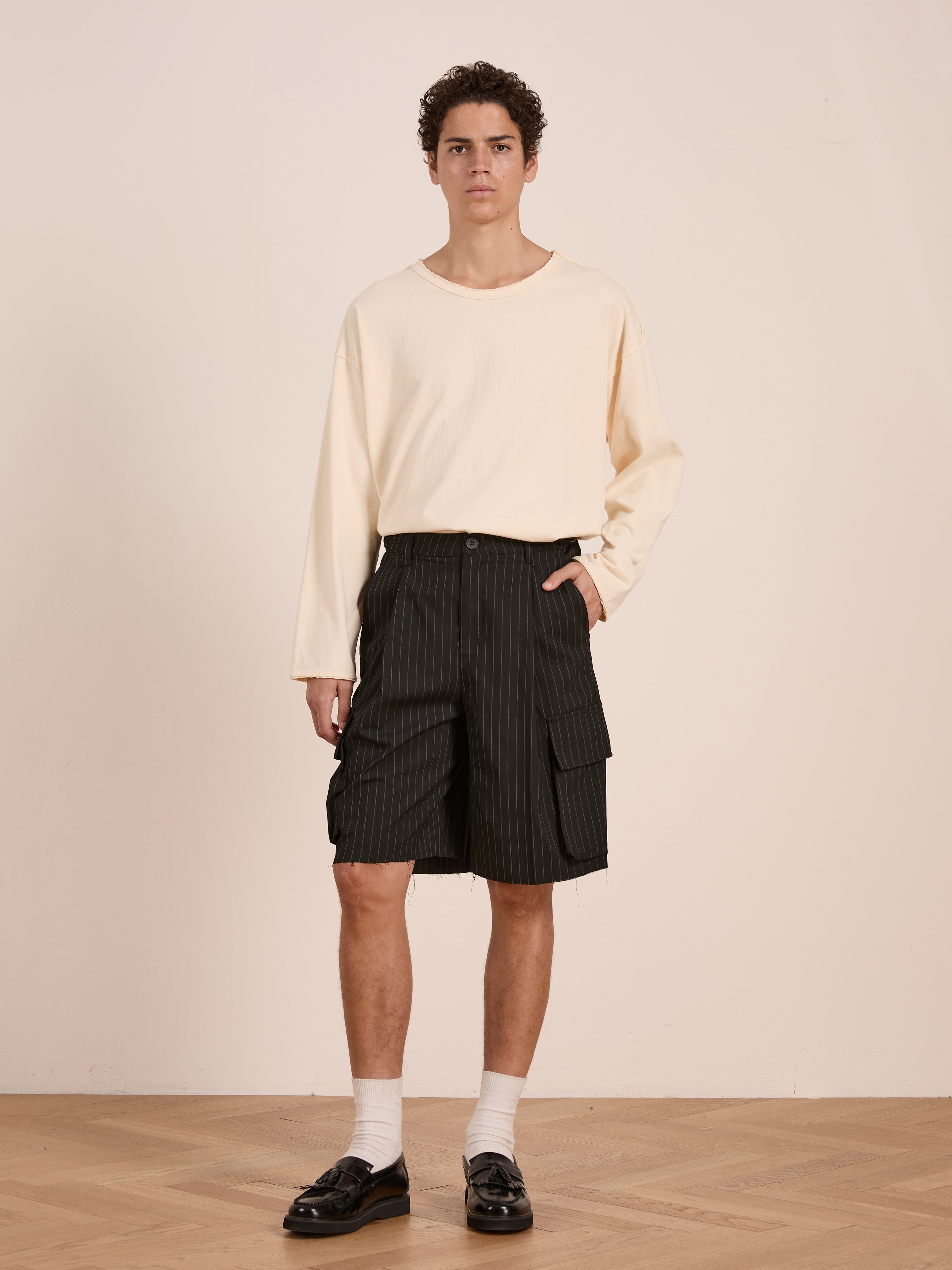A person stands indoors wearing a loose cream long-sleeve shirt and FOUND Hava Pinstripe Pleated Cargo Trouser Shorts, styled with white socks and black loafers against a plain background.