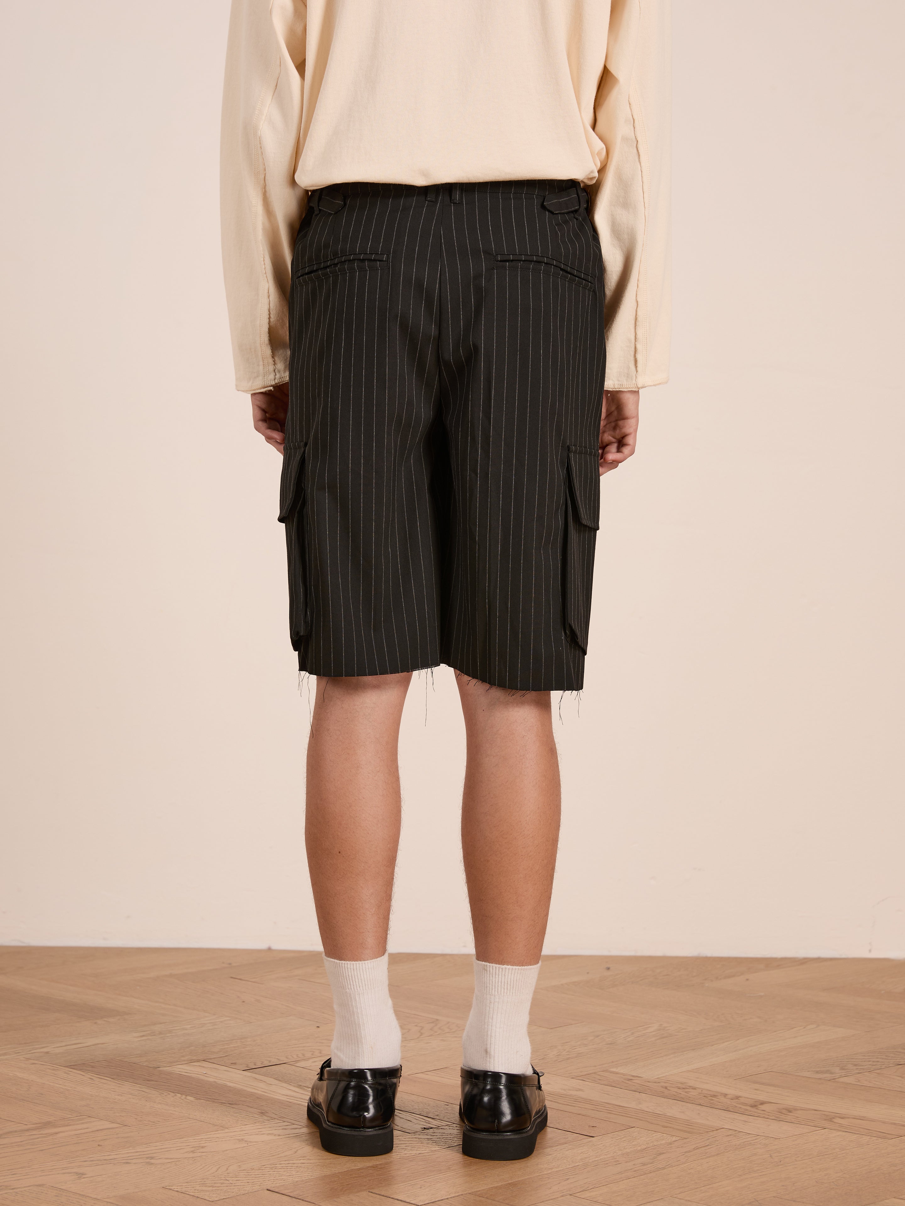 A person stands indoors on a wooden floor, seen from behind, wearing FOUND's Hava Pinstripe Pleated Cargo Trouser Shorts, a beige long-sleeve shirt, white socks, and black shoes.