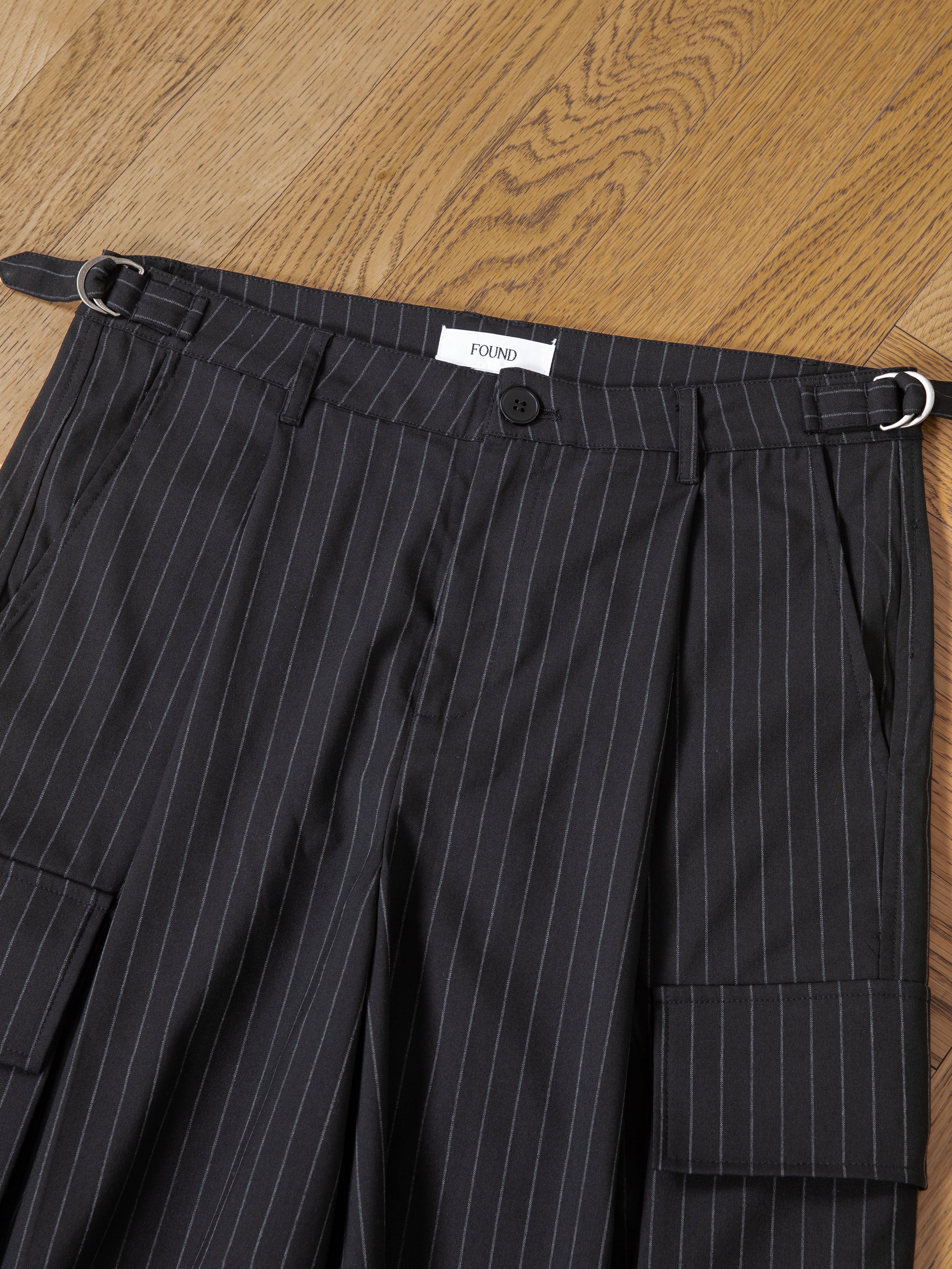 The FOUND Hava Pinstripe Pleated Cargo Trouser Shorts feature a button and zipper closure, belt loops, metal side rings, flap pockets, and an interior "FOUND" label. These unisex shorts are laid flat on a wooden floor for display.