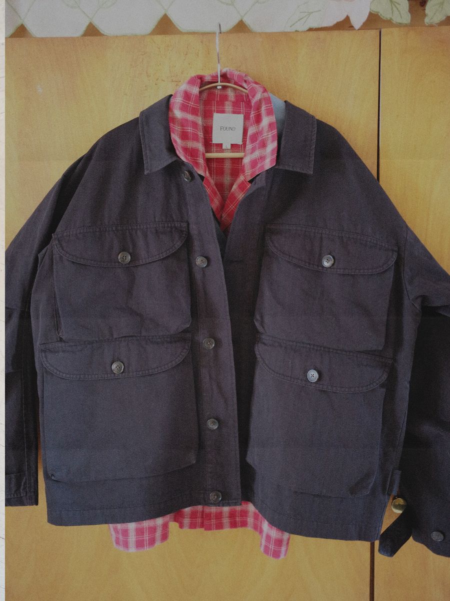 A FOUND Utility Herringbone Cargo Jacket with a vintage vibe is draped over a pink plaid shirt on a hanger, set against a wooden surface.