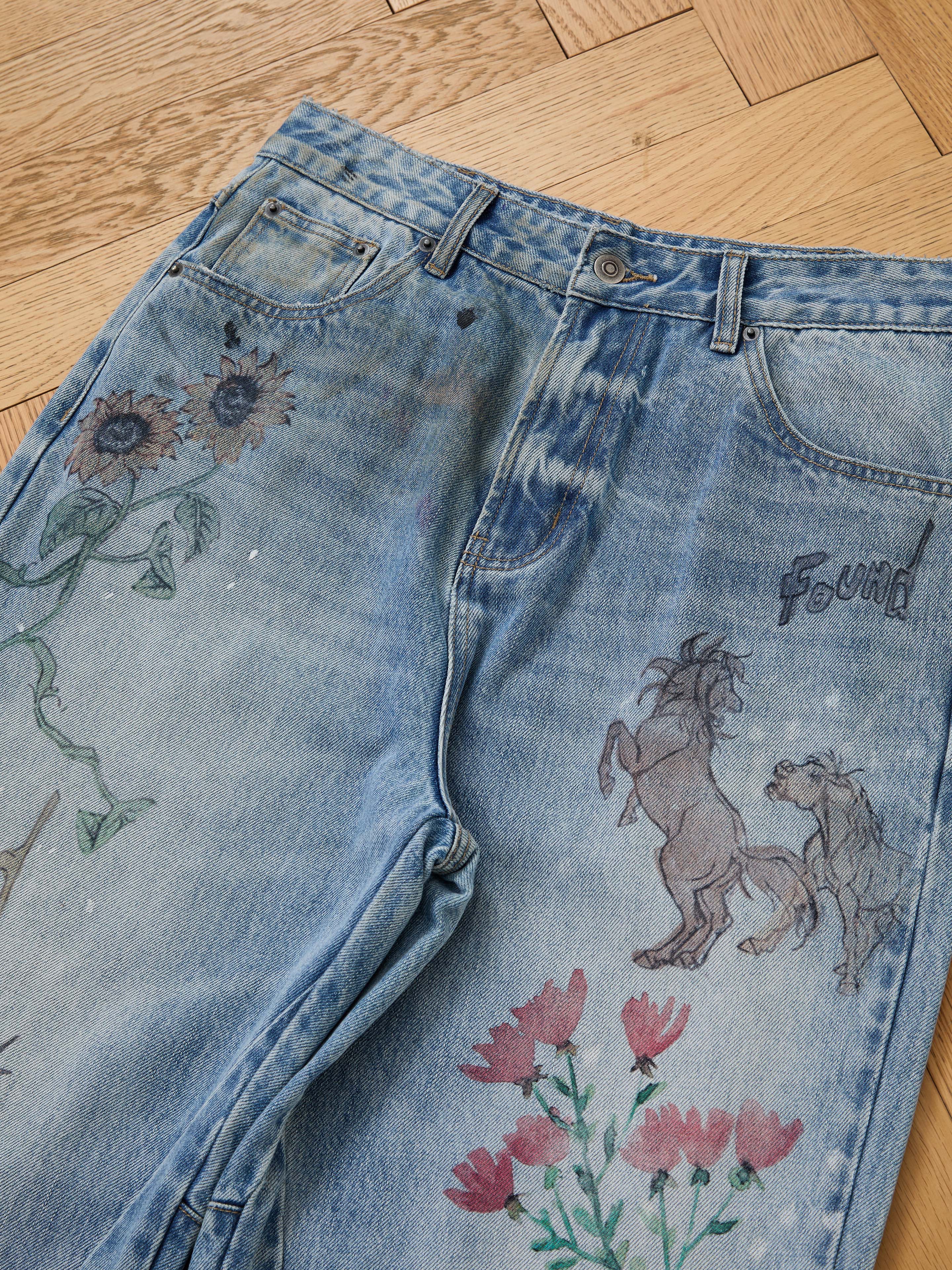 FOUND’s Hand Art Jeans in light blue denim feature unique hand-drawn-style illustrations—sunflowers, pink flowers, two horses, and "found" by the pocket—displayed on a wooden floor.