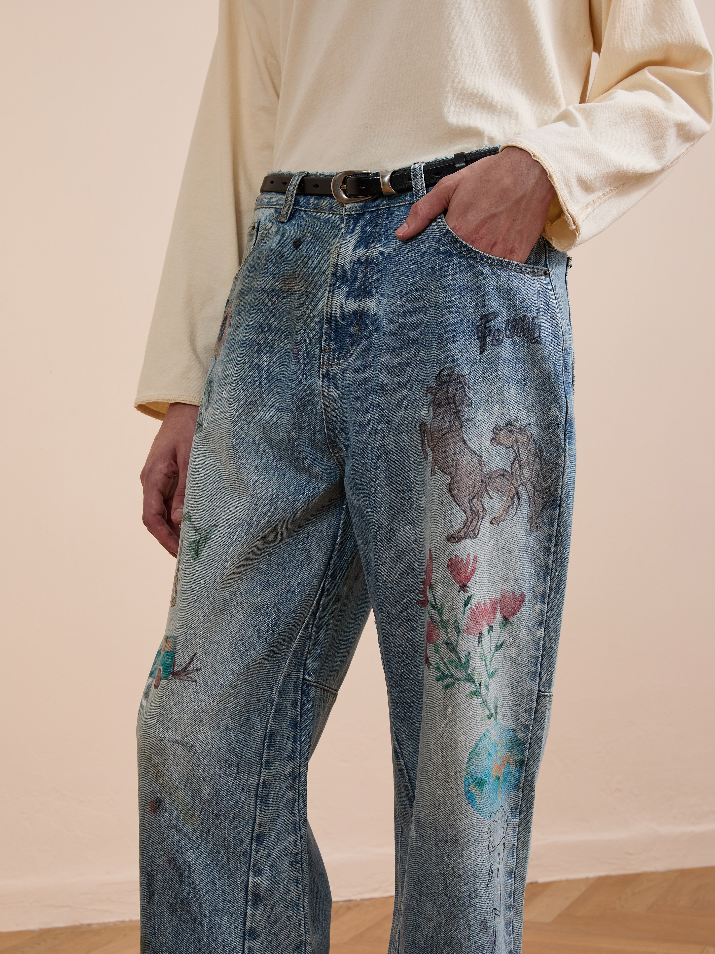 A person wears FOUND Hand Art Jeans—light blue denim with hand-drawn style animals, flowers, and globe designs—paired with a light long-sleeve shirt and a black belt.
